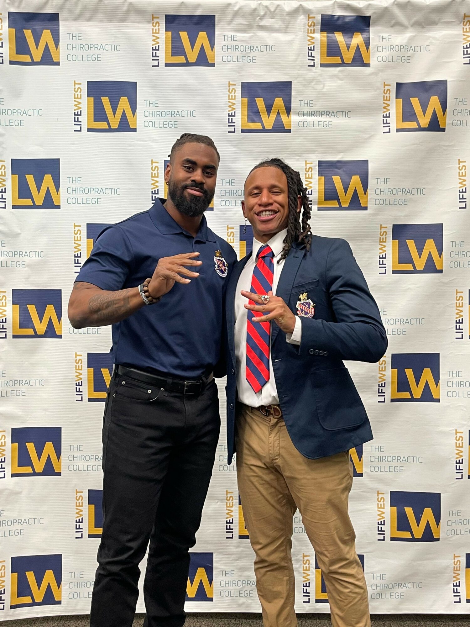 Two men standing in front of a backdrop with a logo, one in a suit and the other in a uniform, smiling and posing.