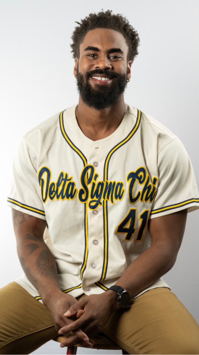Smiling man with curly hair and beard wearing a baseball jersey with text and number, sitting with hands clasped.