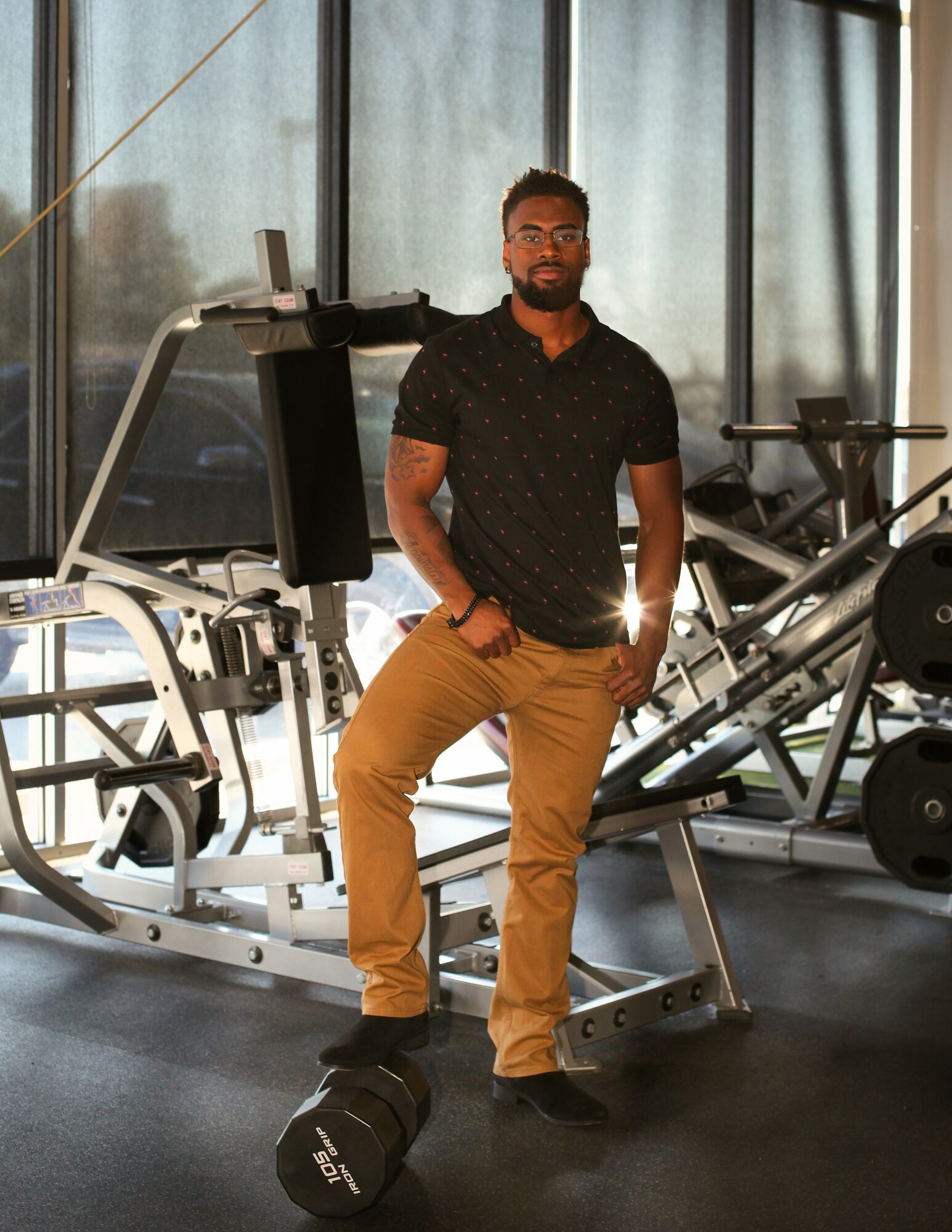 Man standing in gym near exercise equipment, hands in pockets, wearing glasses, black shirt, tan pants, black shoes.