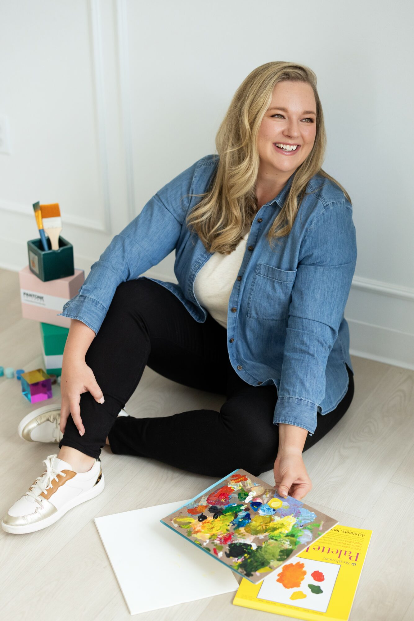 Smiling woman sitting on the floor with art supplies, wearing a denim jacket and white sneakers.