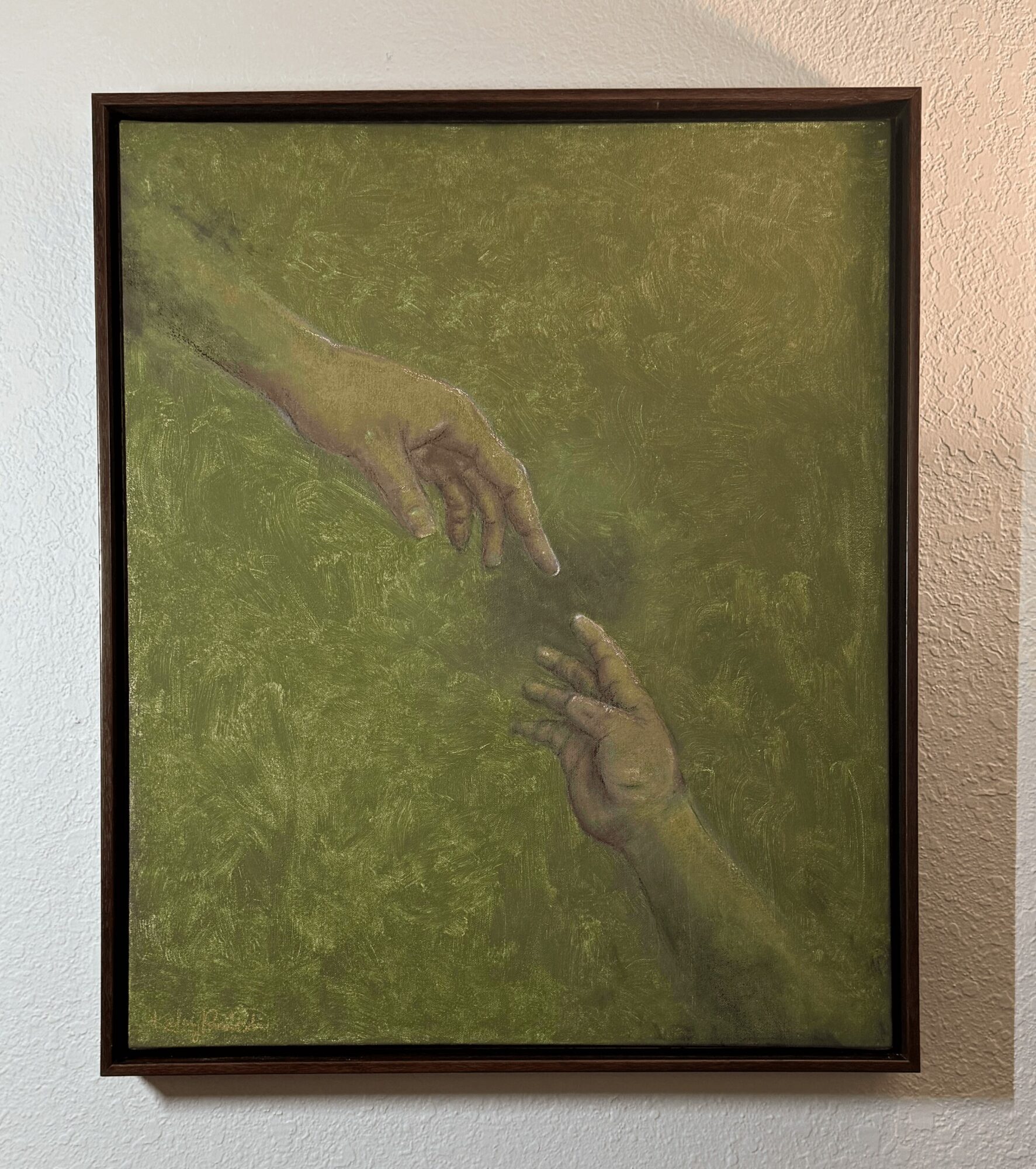 Framed artwork of two hands reaching towards each other against a green background, hanging on a white wall.
