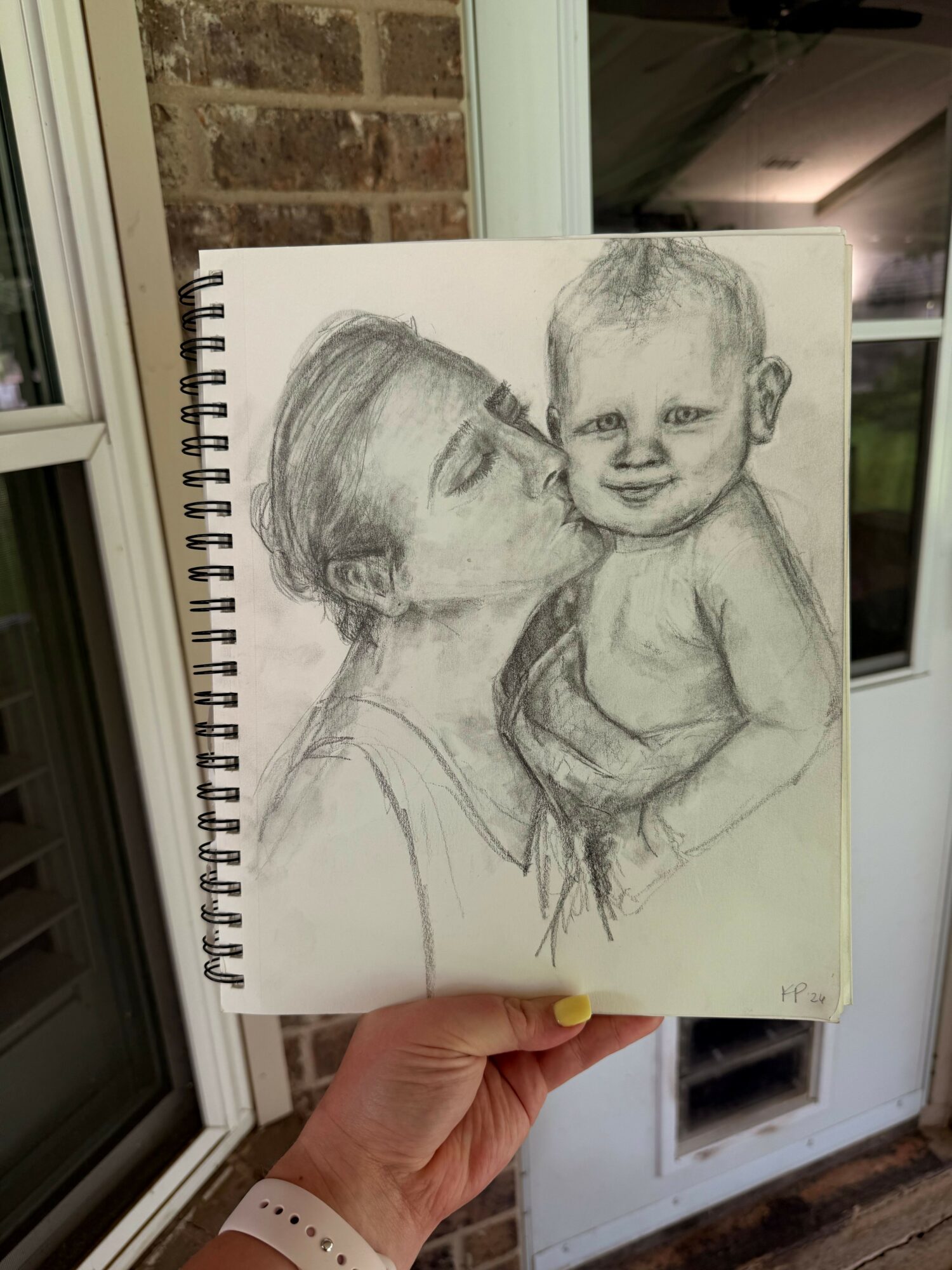 Hand holding a sketchbook with a pencil drawing of two people smiling, outside near a window and brick wall.