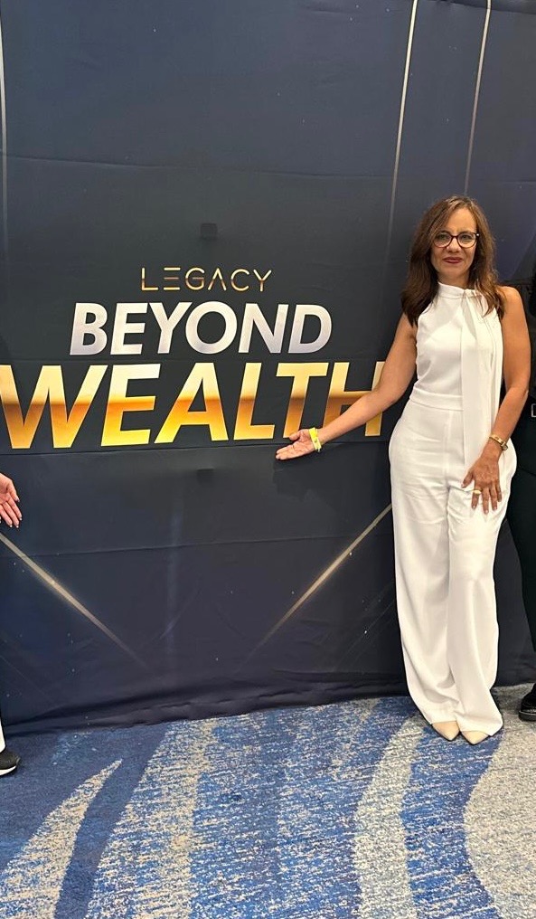 Woman in white pants and top standing next to a sign that reads 'Legacy Beyond Wealth'.
