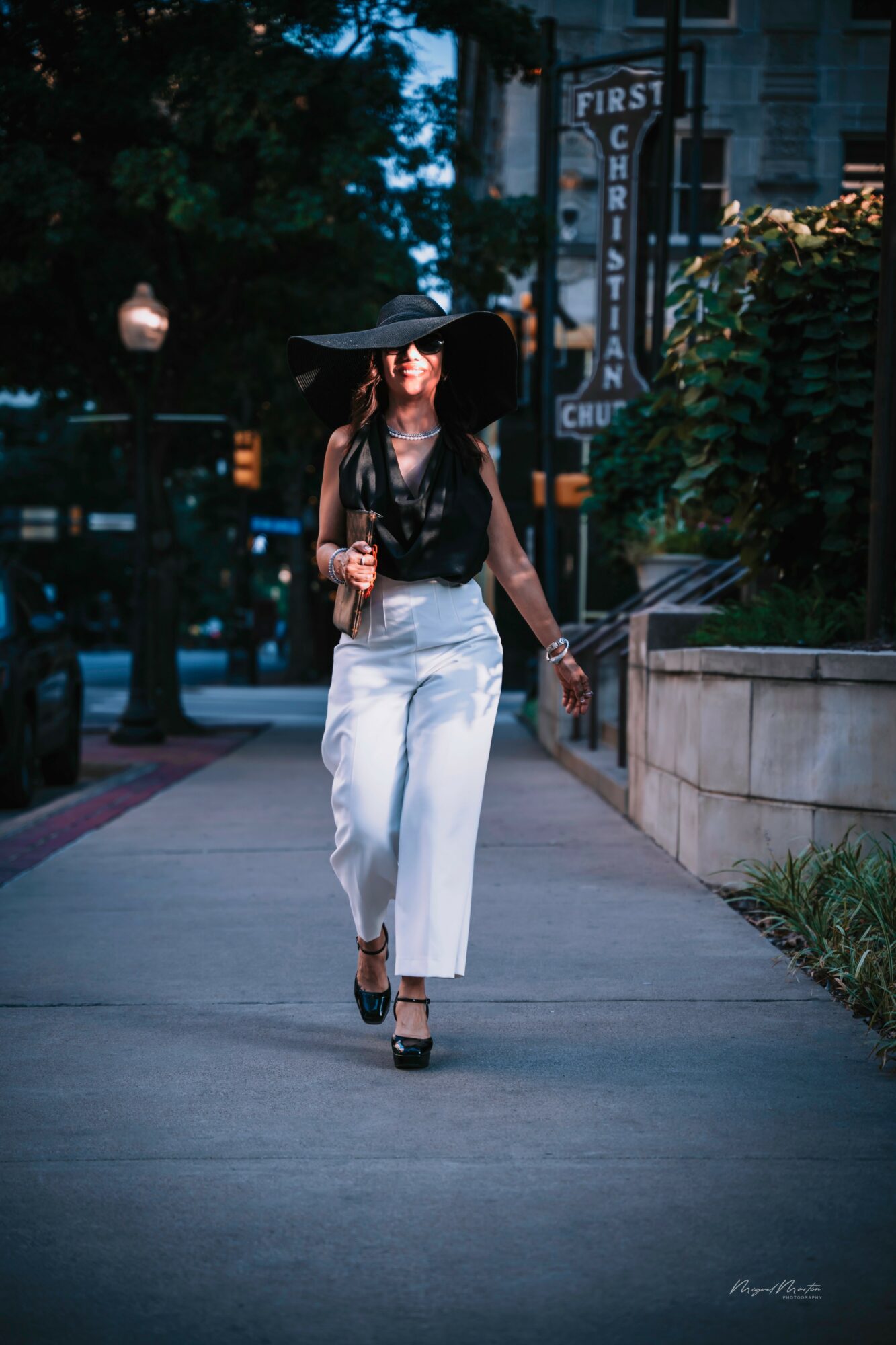 Woman walking on sidewalk holding umbrella, wearing black top and white pants, in an urban setting during evening.