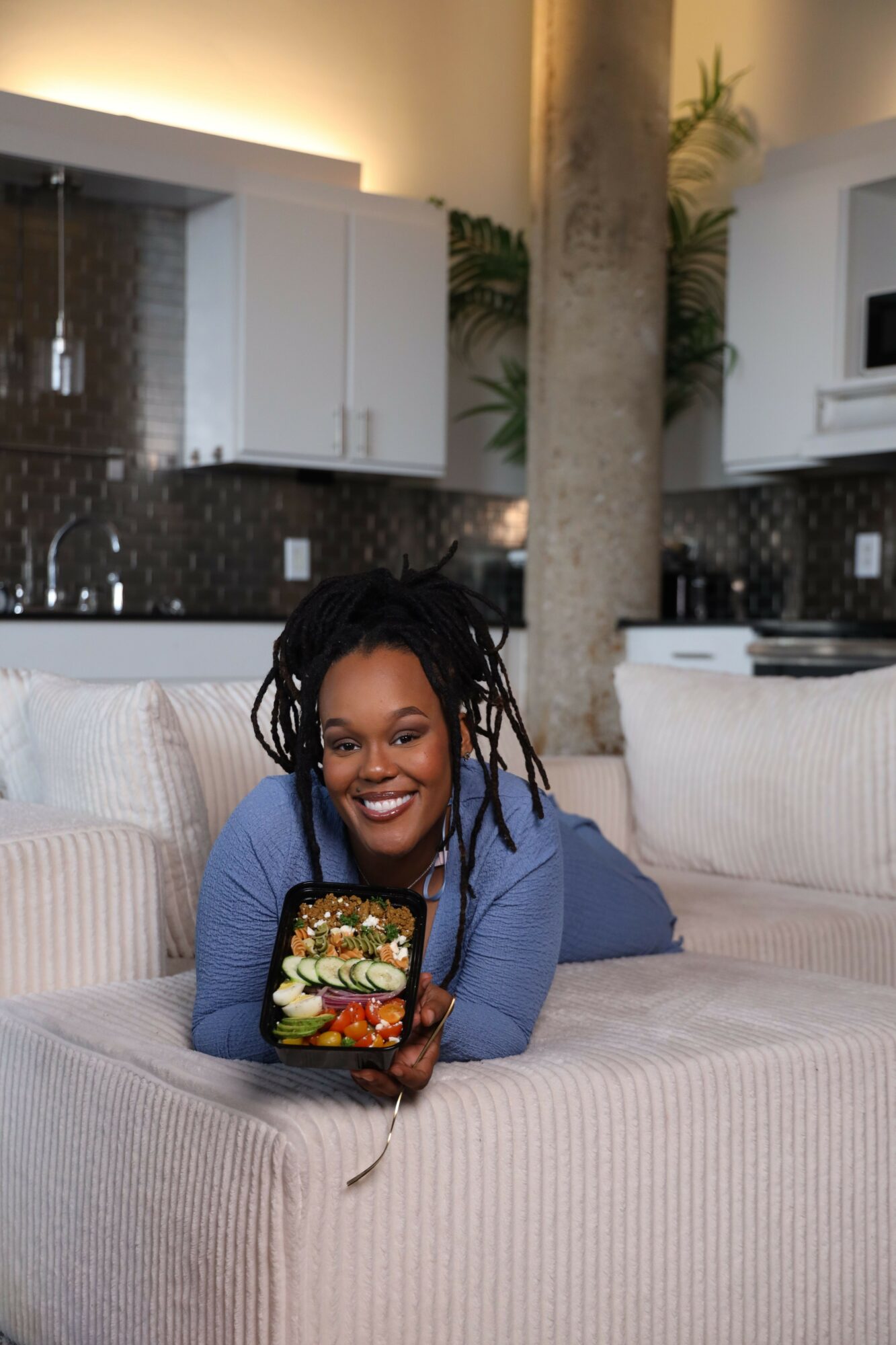 Woman with dreadlocks lying on a sofa, holding a phone with a floral case, smiling at the camera in a modern living room.
