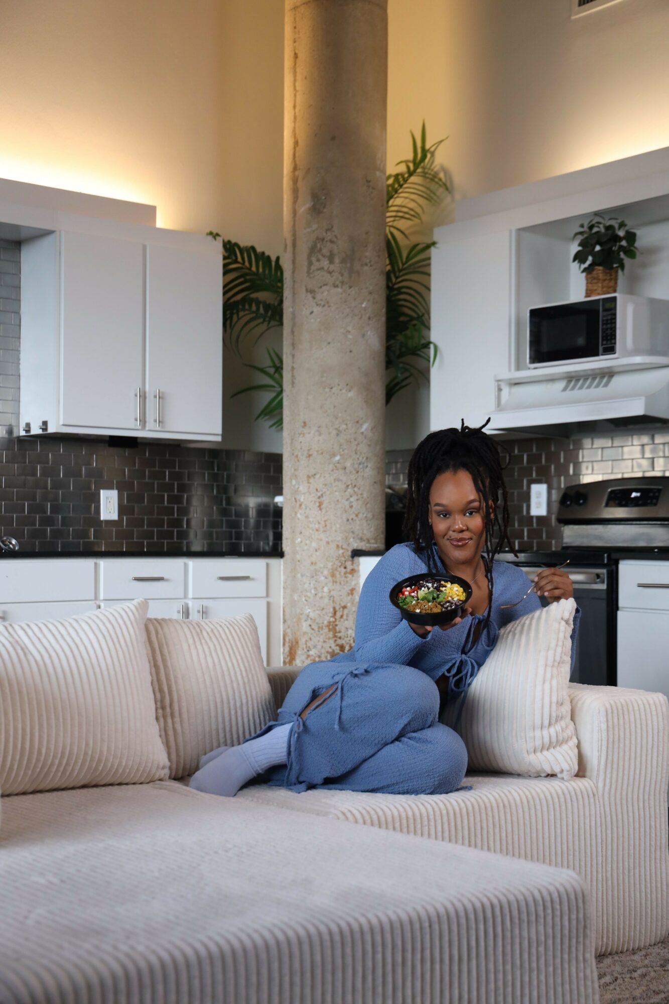 Woman sitting on a sofa in a modern living room, holding a phone, with a tree and kitchen in background.