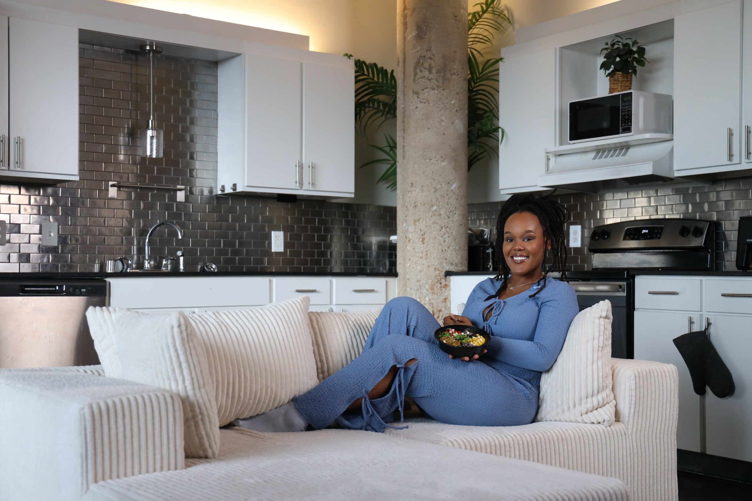 Woman in blue outfit sitting on a white couch in a modern kitchen, smiling, holding a bowl.