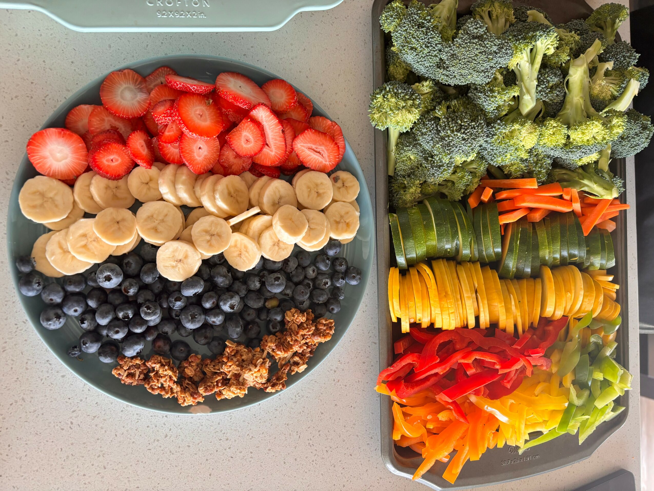 Plate with sliced strawberries, bananas, blueberries, and granola; side tray with broccoli, carrots, yellow and red bell peppers, and cucumber slices.