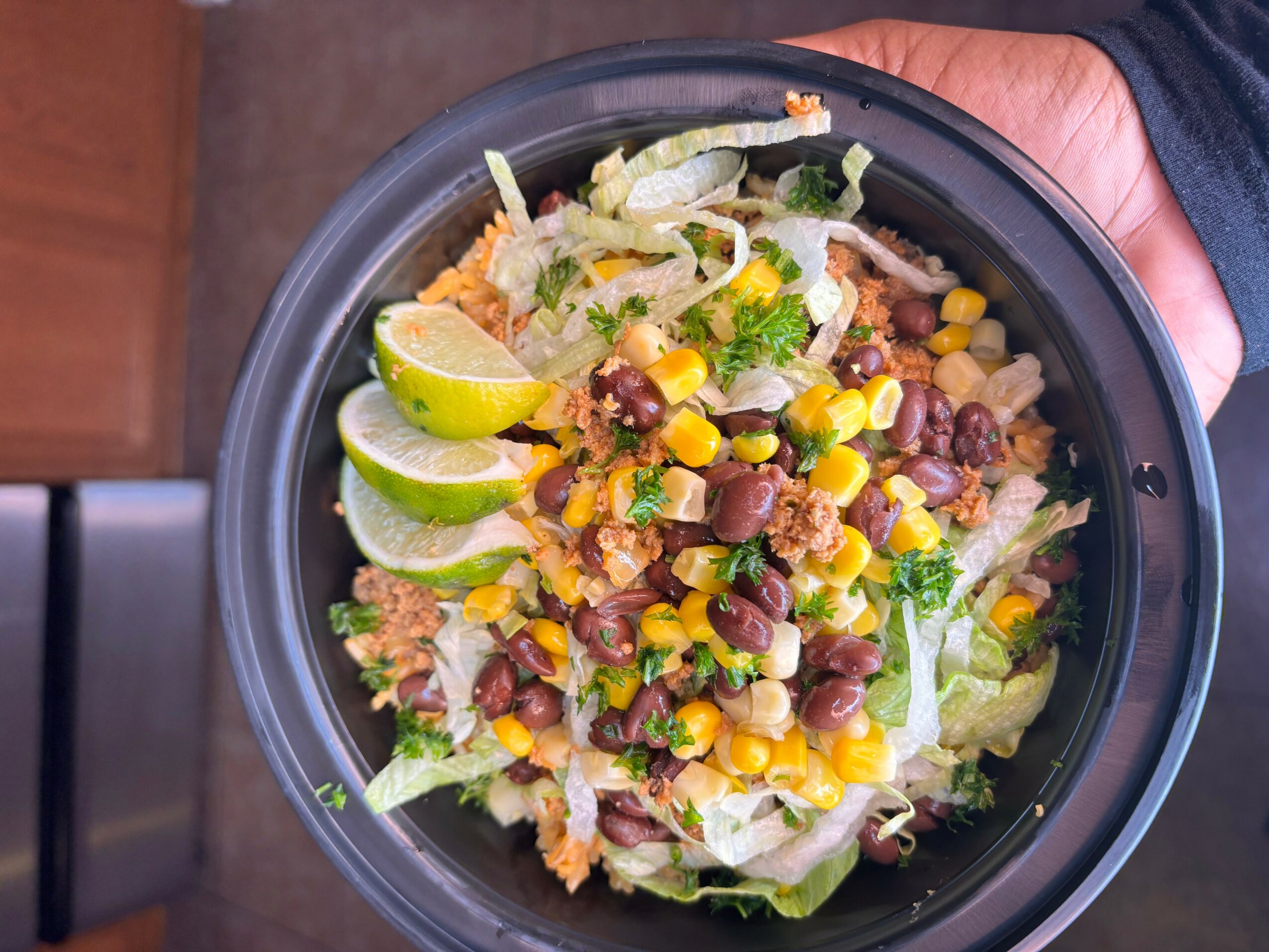 Bowl of salad with lettuce, corn, black beans, lime wedges, and shredded cheese, held by a person.
