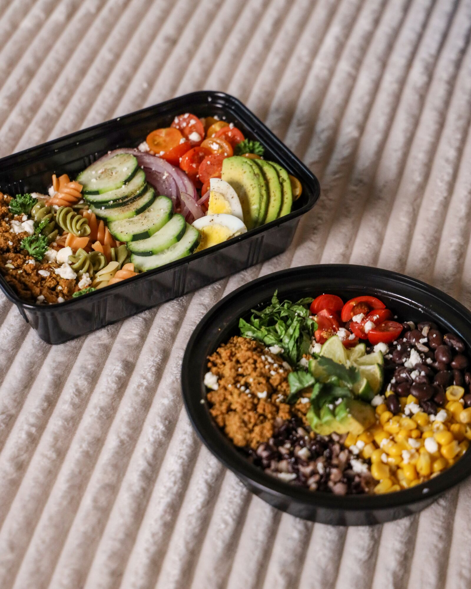 Two black containers with salads and toppings on a beige striped fabric, one with cucumbers, cherry tomatoes, and other vegetables, the other with grains, corn, and greens.