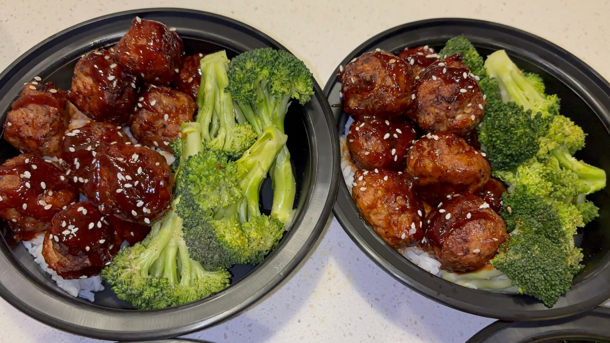 Two black bowls with broccoli and glazed meatballs, sprinkled with sesame seeds, on a white surface.