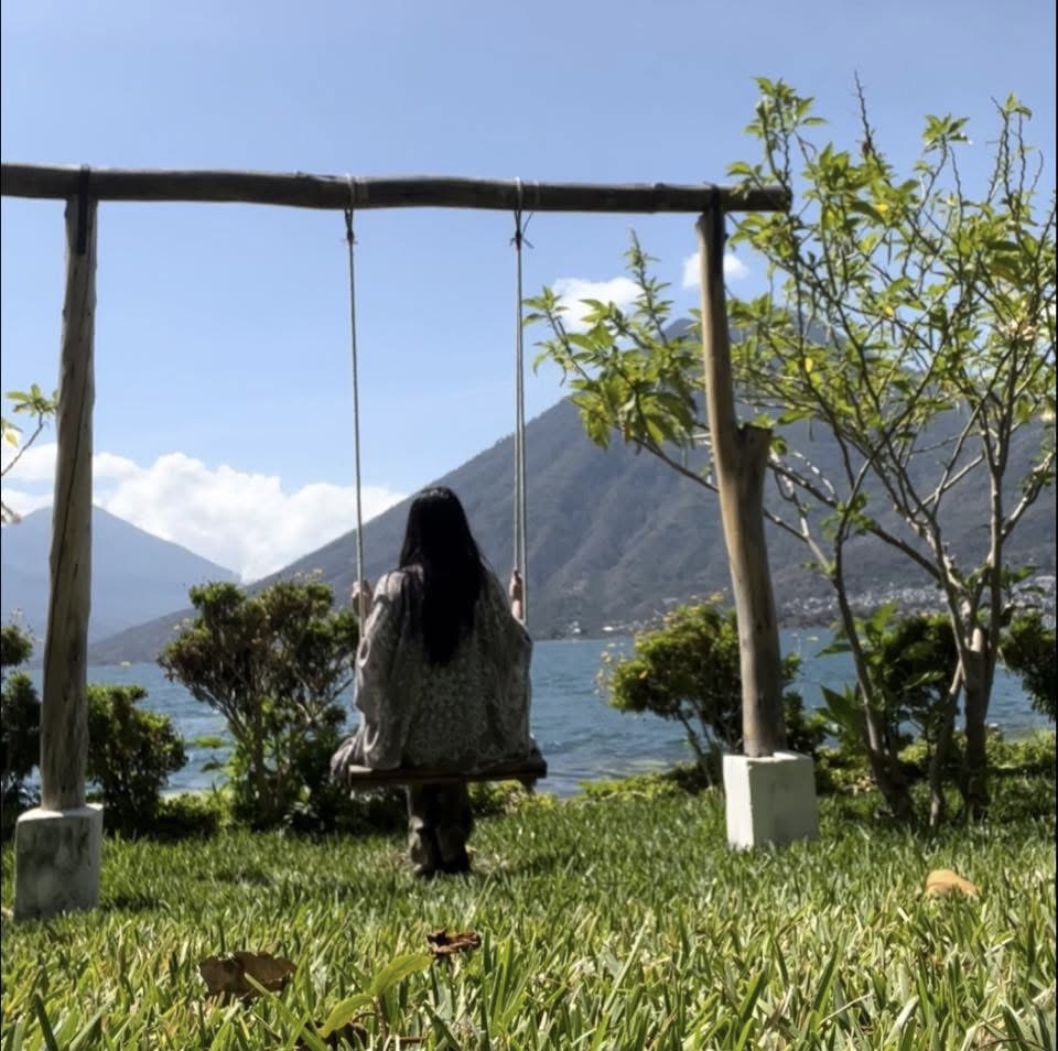 Person sitting on a swing facing a lake with mountains and trees in the background.