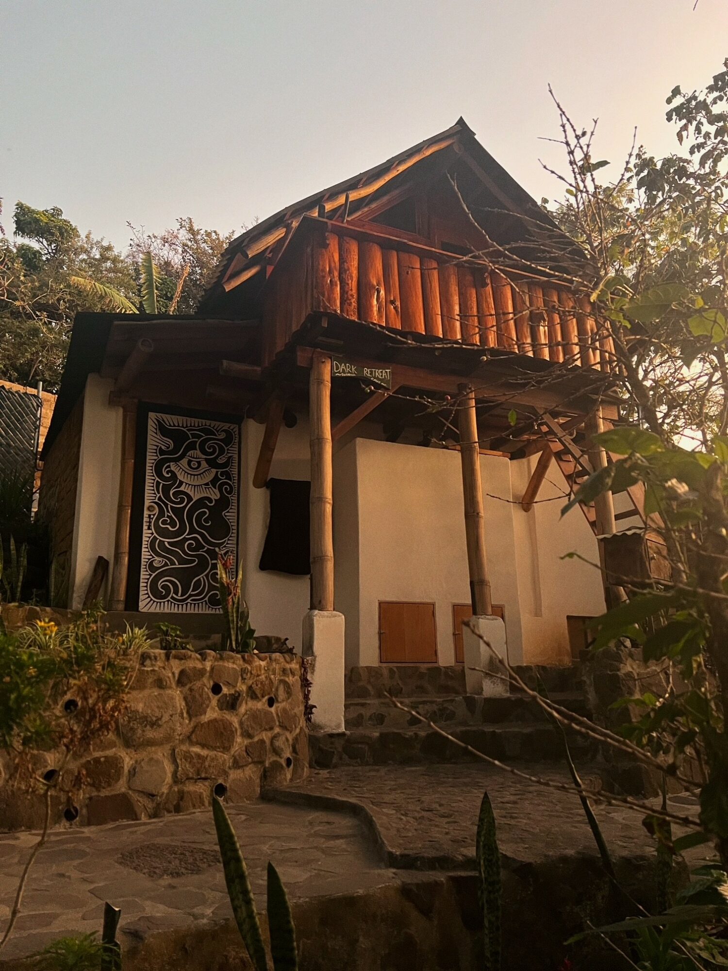 Traditional wooden house with a steep roof, elevated on stilts, surrounded by greenery and stone steps.