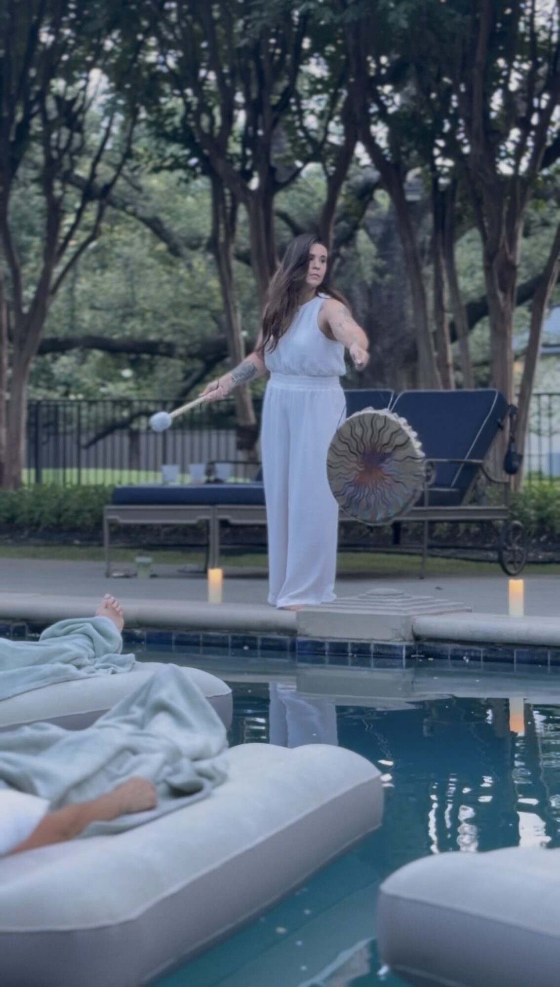 Woman in white pants and top standing near poolside lounge chair with umbrella, trees in background.