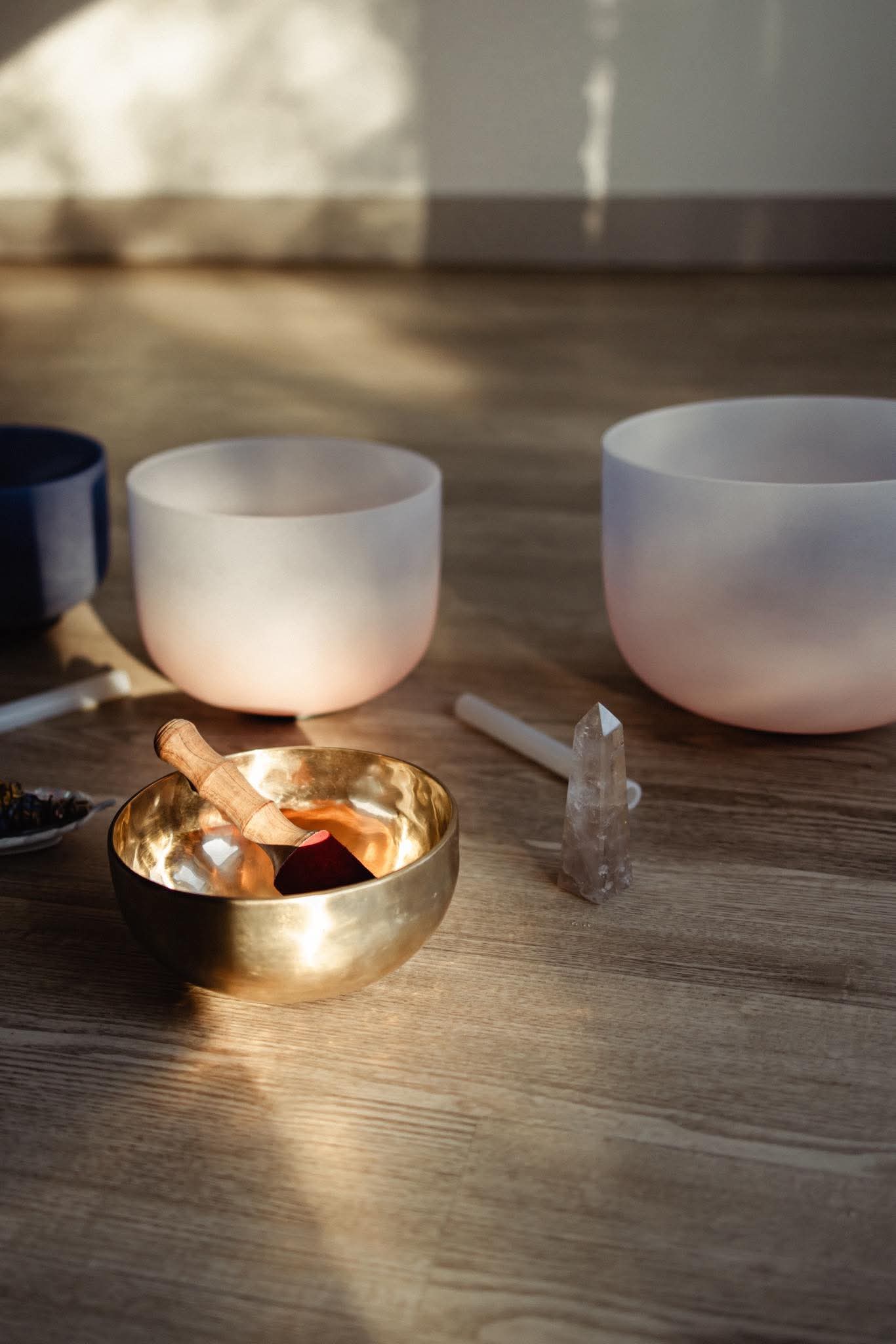 Three singing bowls of different sizes on a wooden surface with sunlight and shadows, and a small crystal.