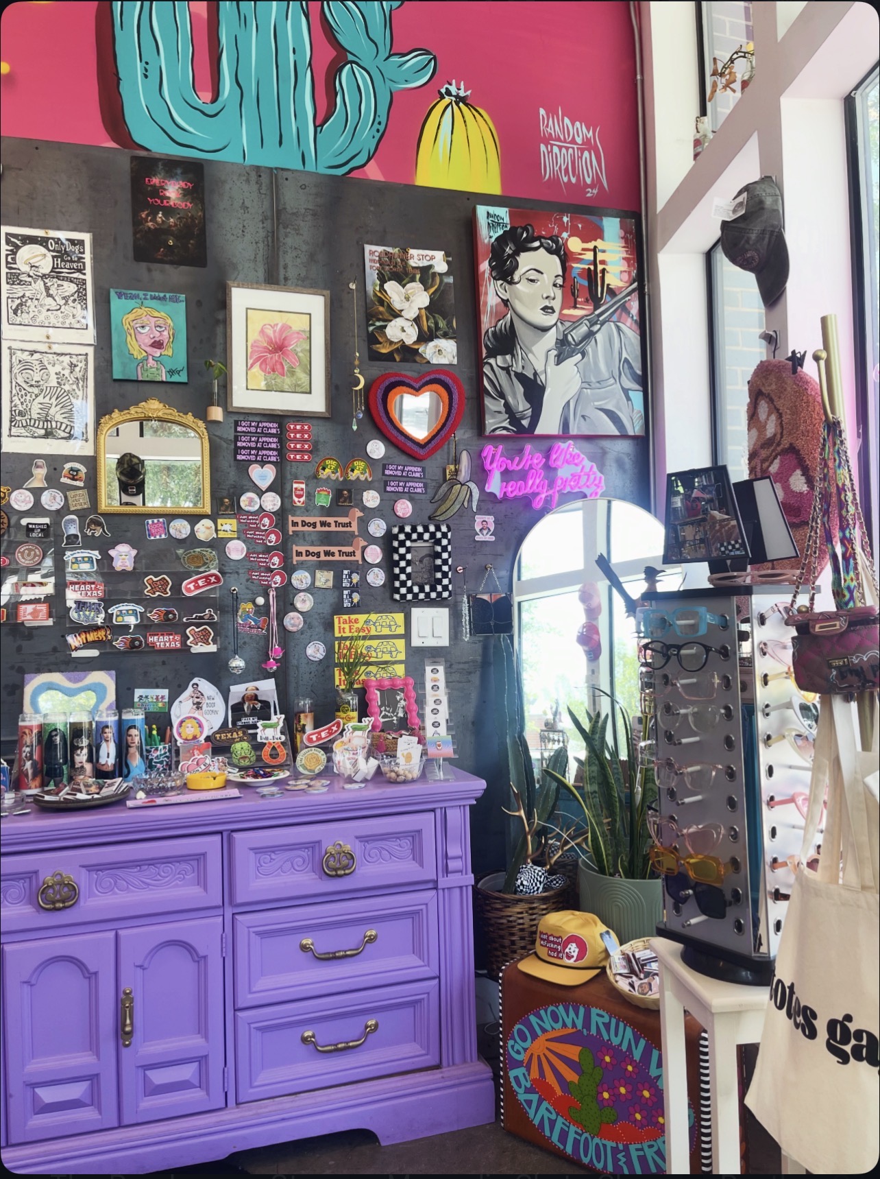 Display of colorful artwork, pins, and accessories on a black wall with a purple dresser below, in a bright room.