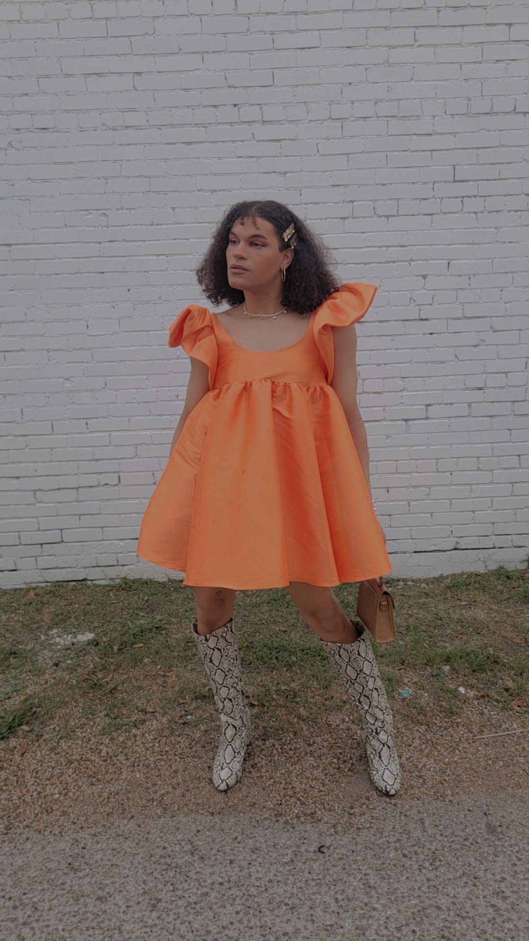 Young woman in orange dress and patterned boots standing outdoors against a white brick wall.