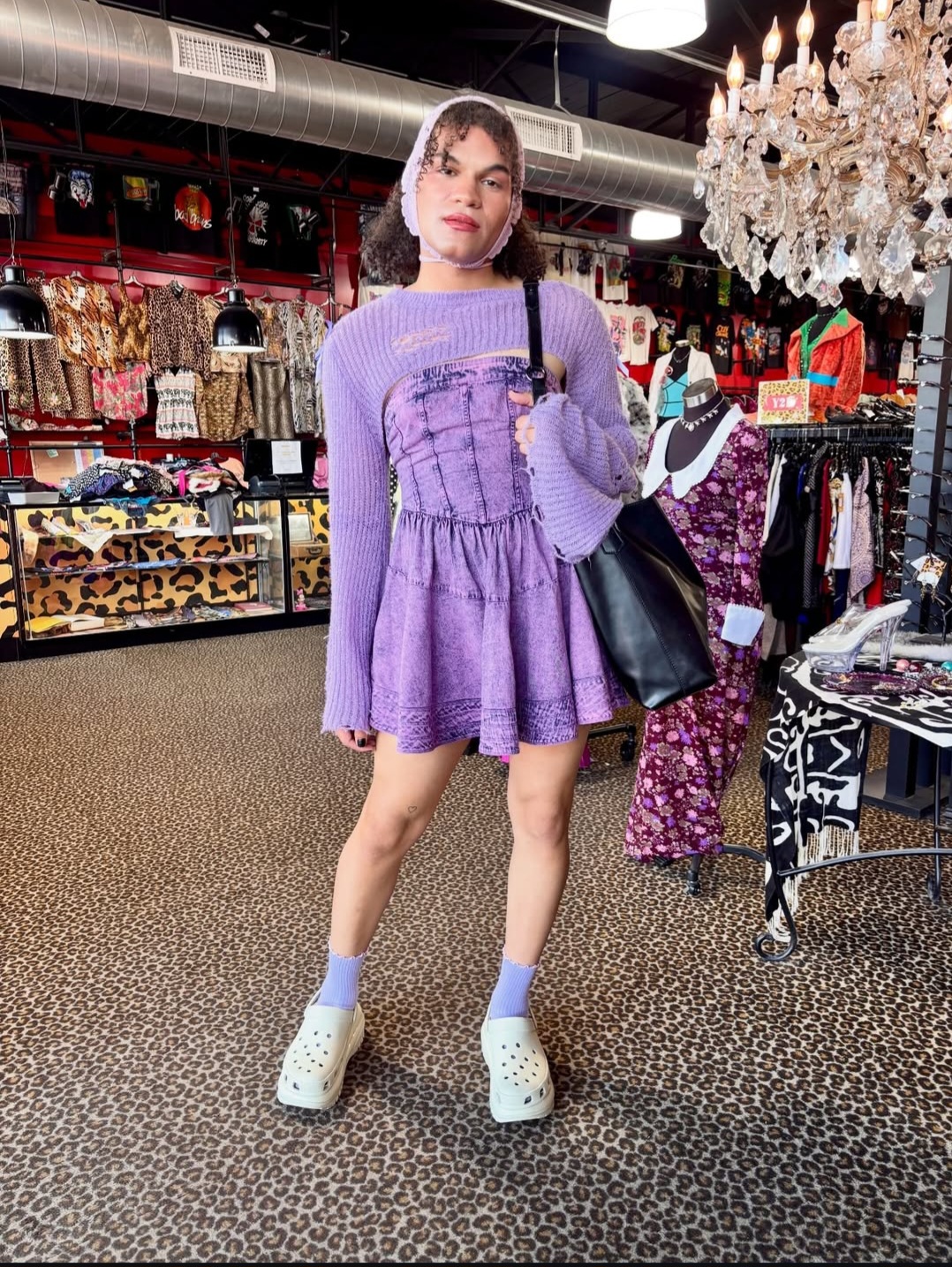 Young woman in purple dress and white shoes stands in store with clothing and accessories, chandelier overhead, leopard print carpet.