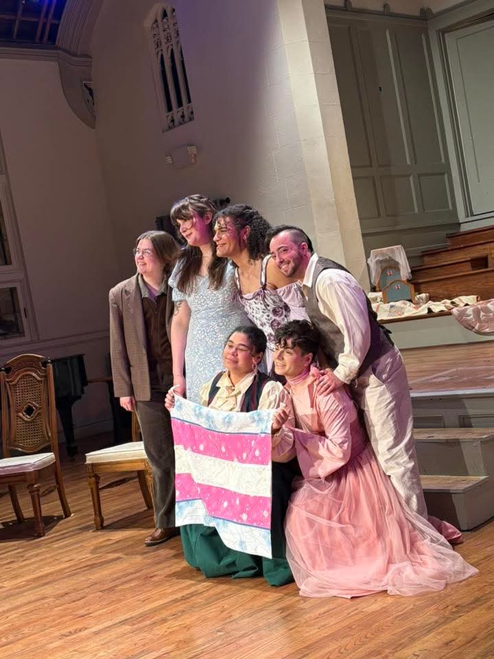 Group of seven people on stage, some holding a pink and white striped cloth, in a room with wooden floor and green walls.