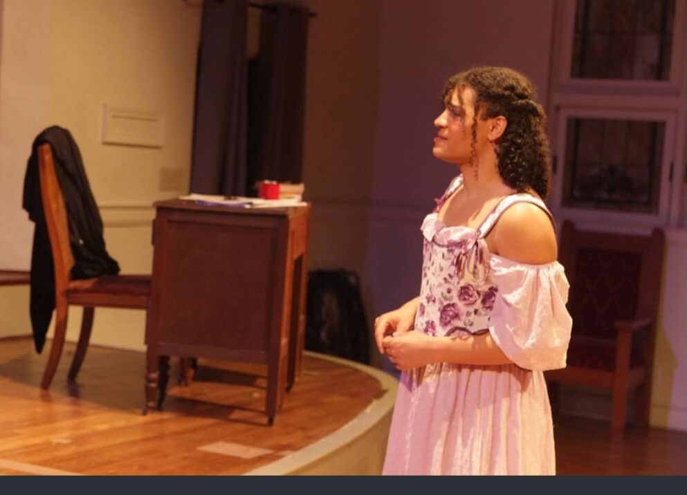 Young woman in a pink dress with off-shoulder sleeves standing on stage, facing right, in a room with furniture.