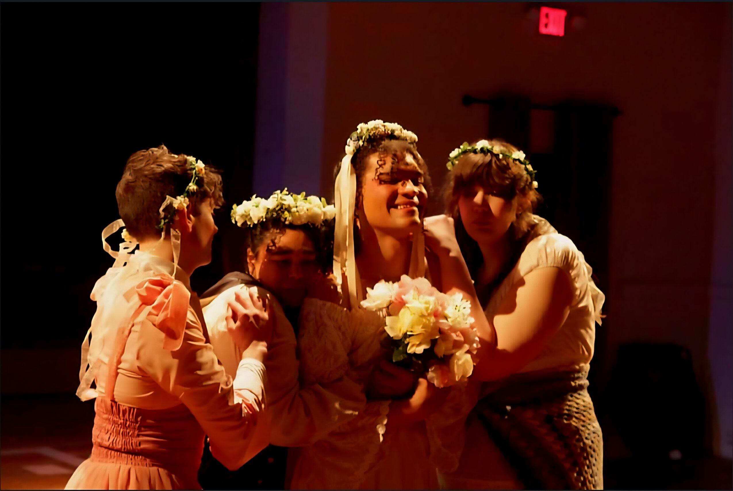 Group of five women wearing floral crowns, holding flowers, smiling in a dimly lit room.