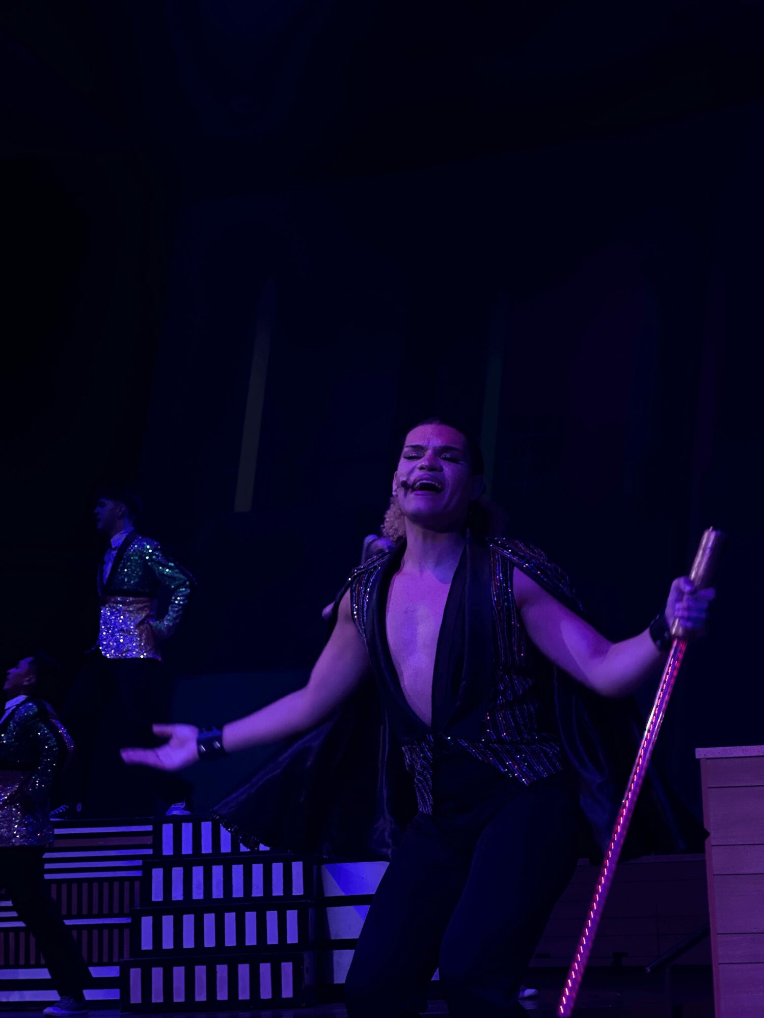 Performer singing on stage with a microphone stand, wearing a black outfit, in a dark setting.