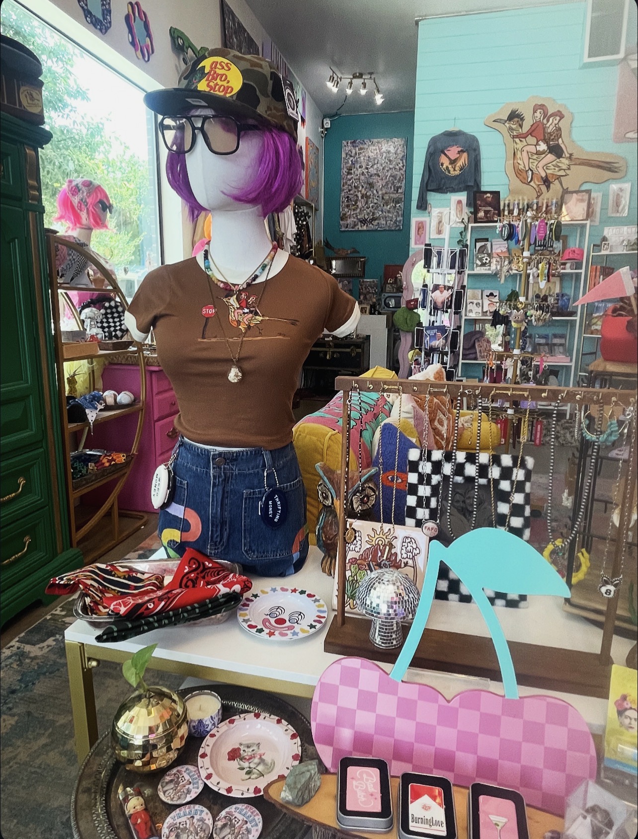 Mannequin with purple hair, glasses, brown shirt, and jeans in a colorful store with accessories and decor.