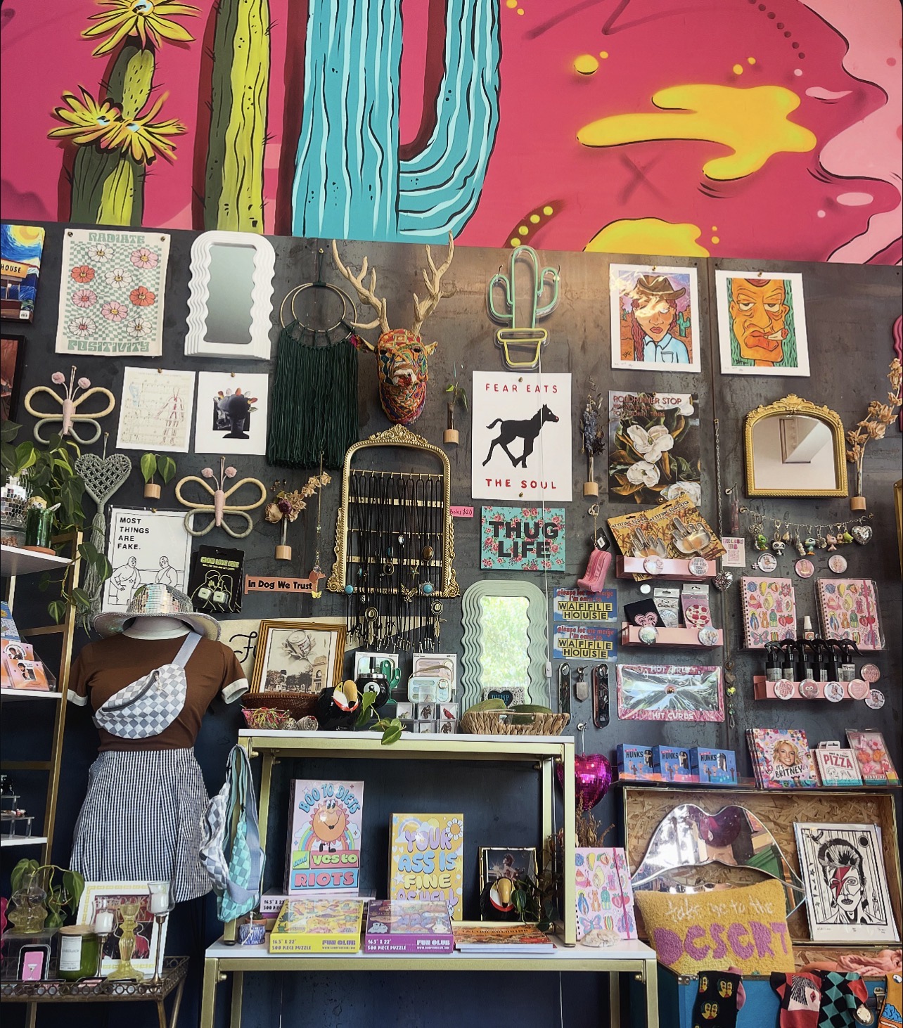 Display of colorful artwork, jewelry, and accessories with a person browsing, set against a decorated wall with various framed pieces.