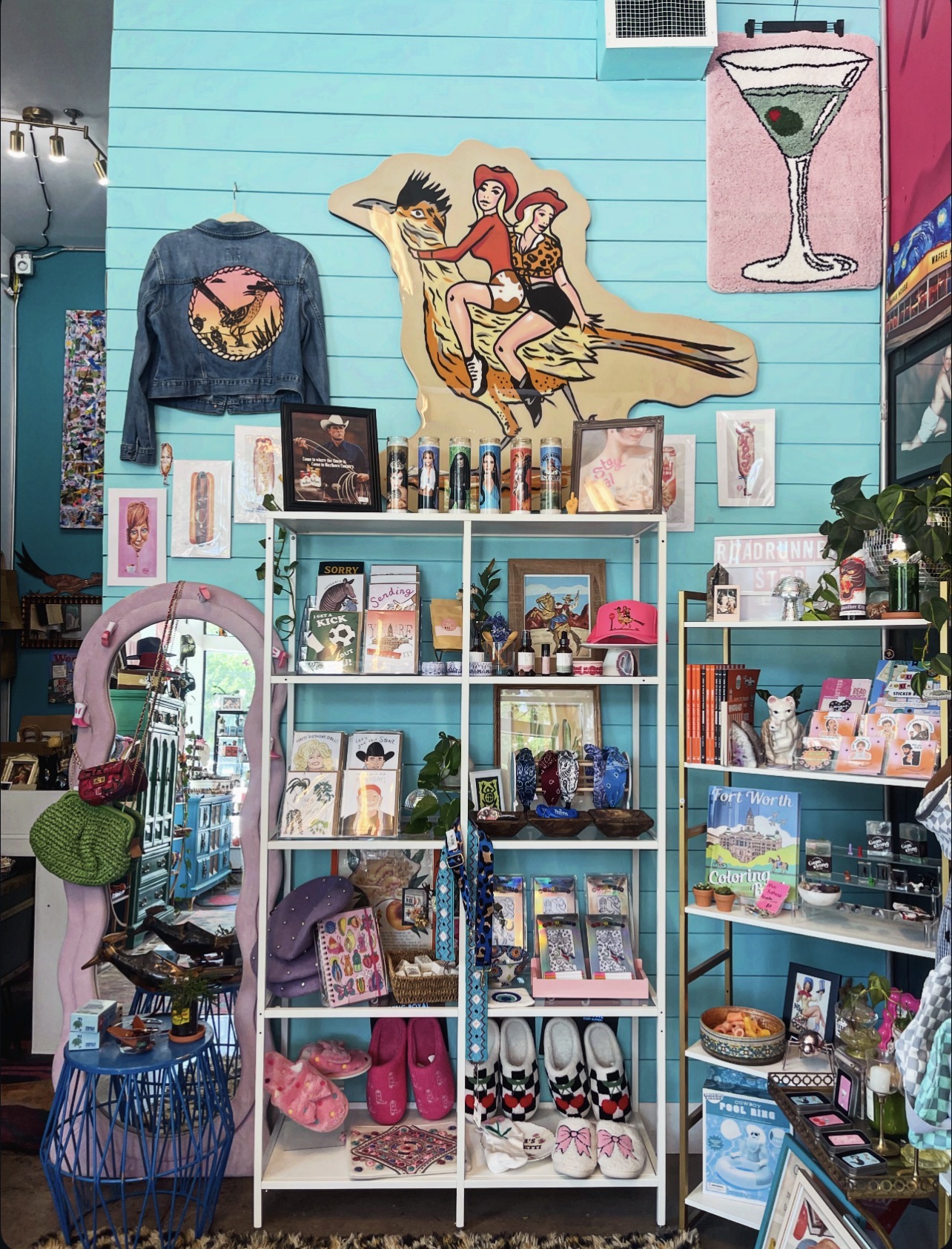 Display of clothing, accessories, and artwork against a blue wall, including a jacket, framed pictures, and decorative items.
