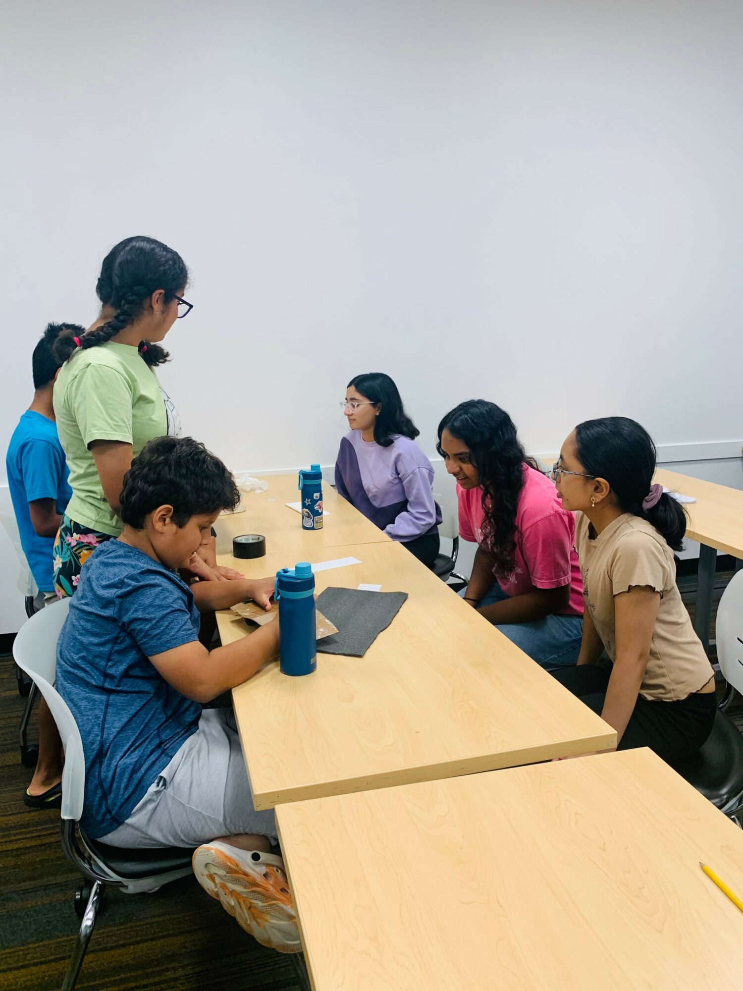 Group of children sitting at a table, engaging in a discussion or activity, with a plain white wall in the background.