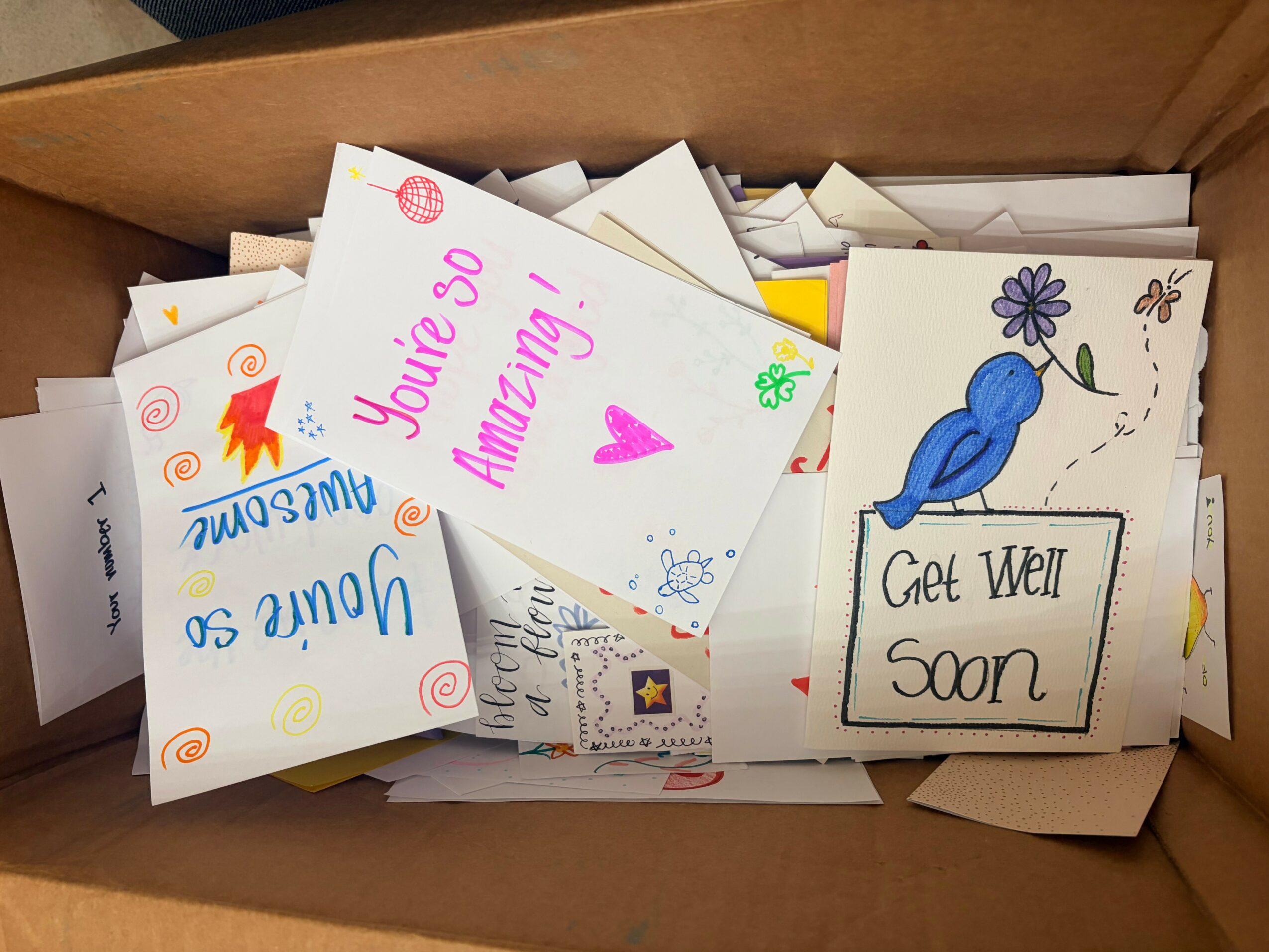 Box filled with colorful greeting cards, including one with a blue bird and a 'Get Well Soon' message, and another with a pink heart and 'You're so Amazing!' text.