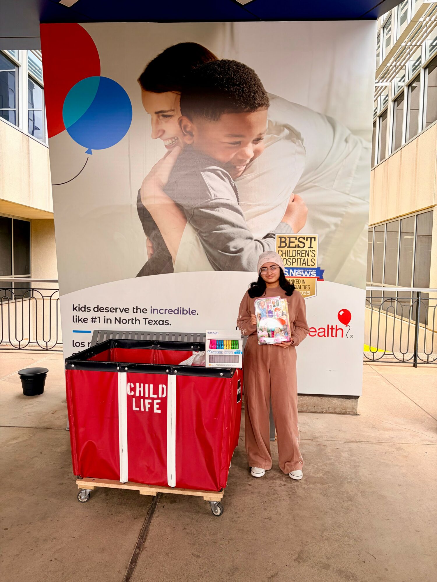 Young girl holding a pamphlet standing next to a red table with a banner, in front of a large poster of two children hugging.