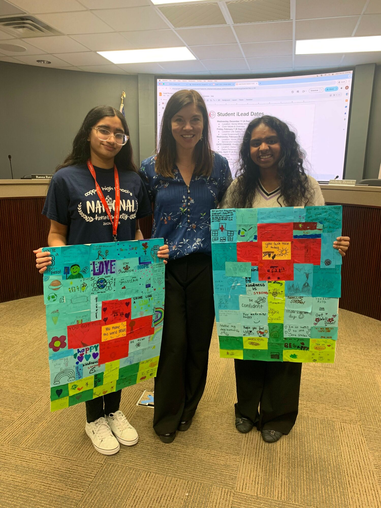 Three women stand in a room holding colorful posters with text and graphics, smiling at the camera.
