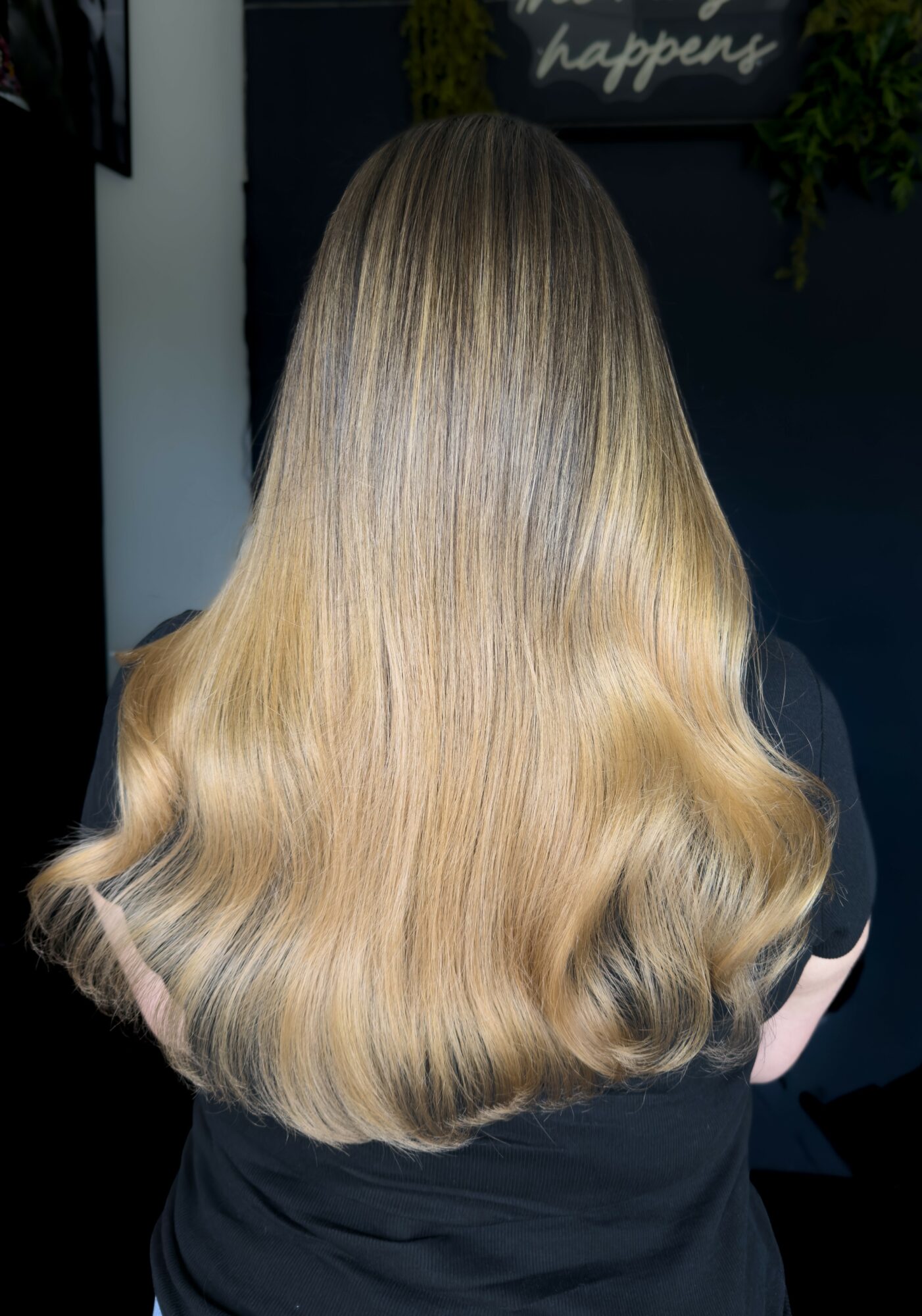 Back of a person with long, wavy blonde hair, standing indoors against a dark background.