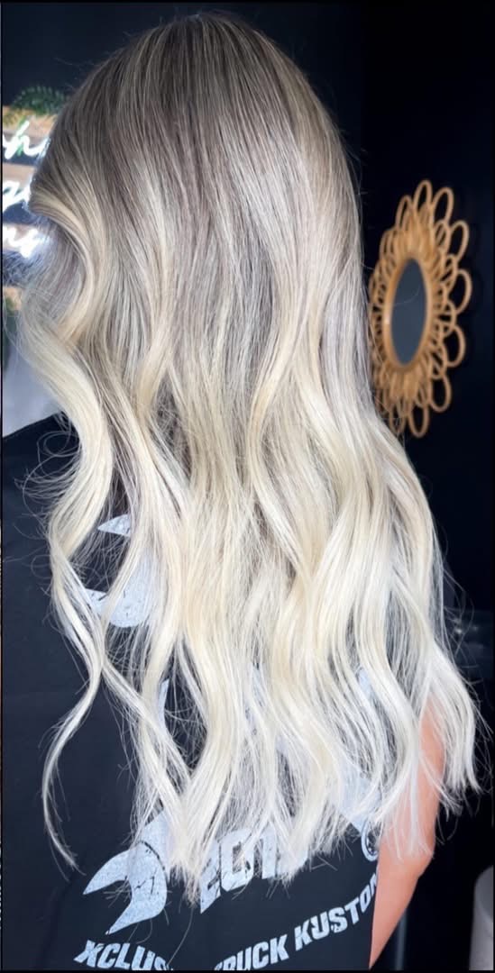 Back of a person with long, wavy, blonde hair, standing in front of a mirror with a decorative frame.