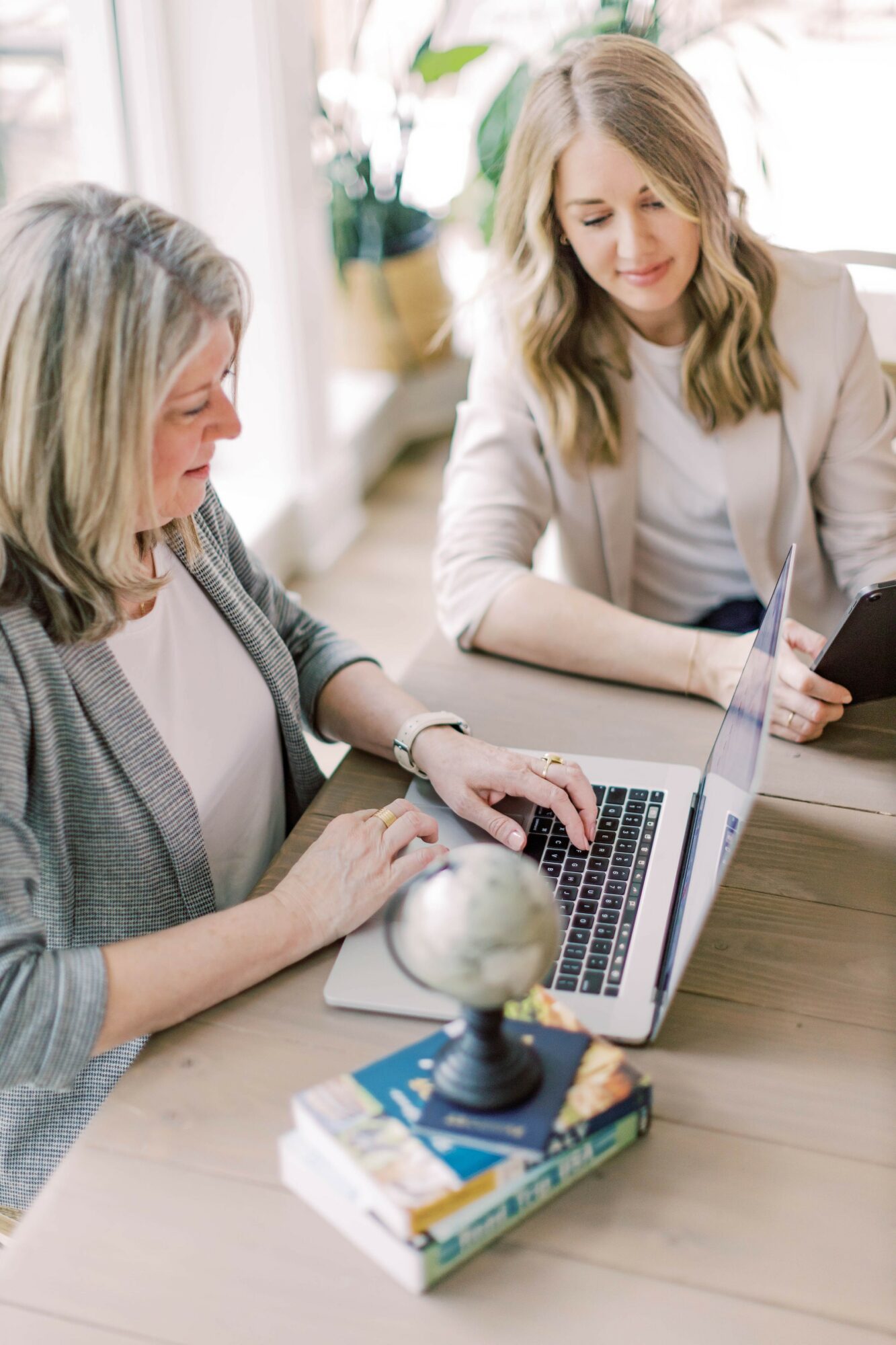 Two women sit at a table with a laptop, globe, and books, engaging in a discussion in a bright room.