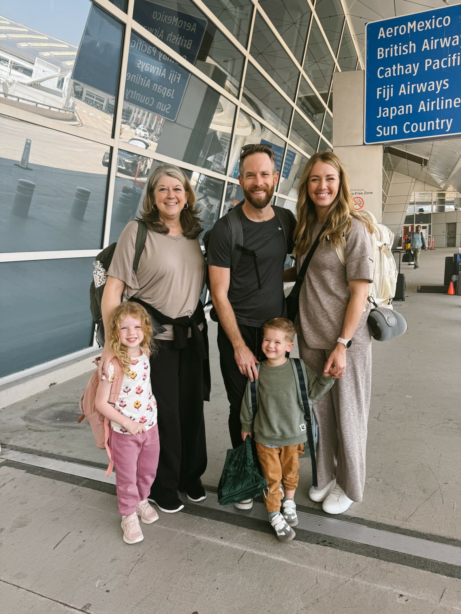Group of five people, two children, standing outside an airport terminal with glass windows and a blue sign, smiling.