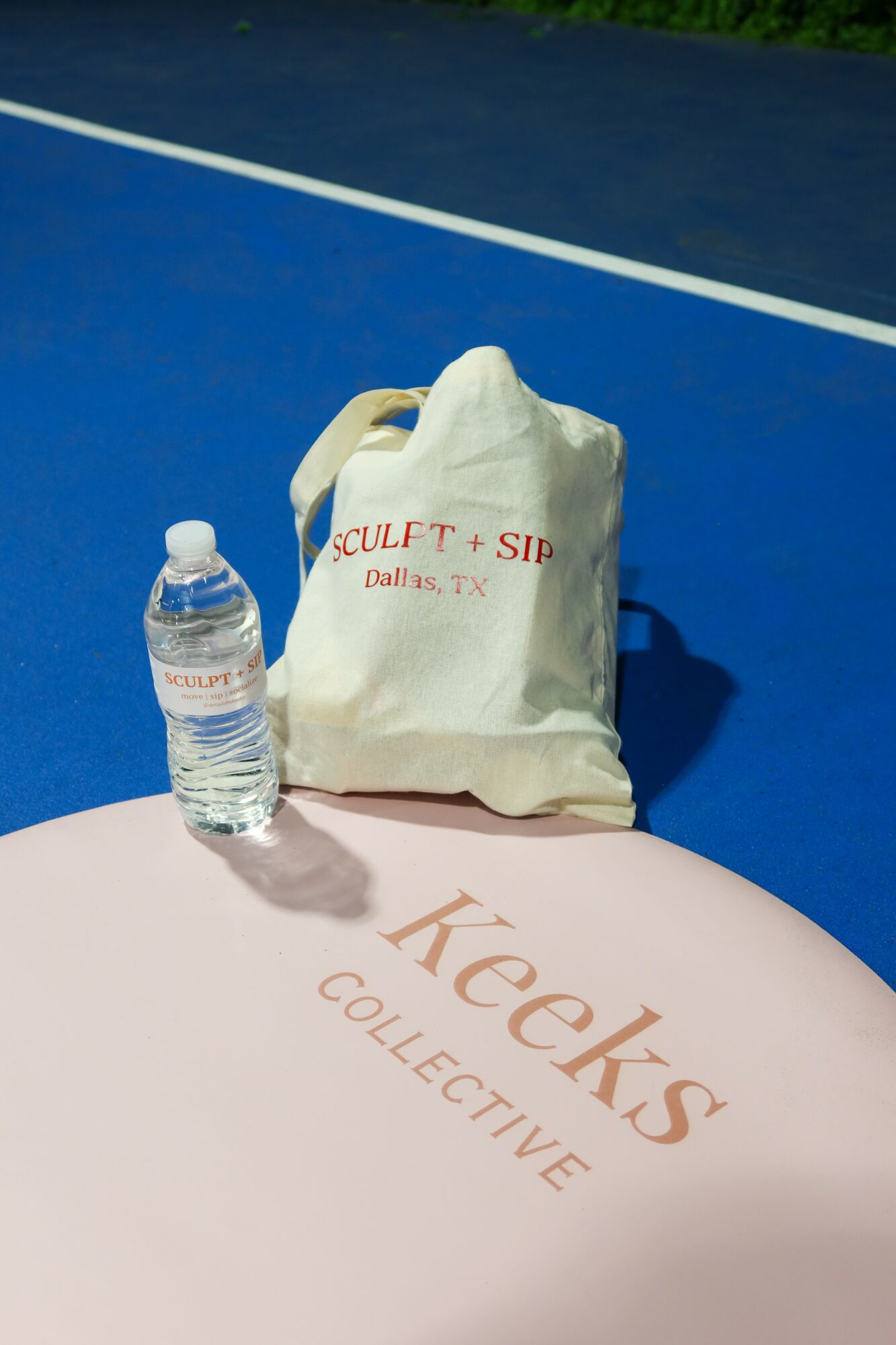 A plastic water bottle and a paper bag on a tennis court with a blue surface and white lines, shadow visible.