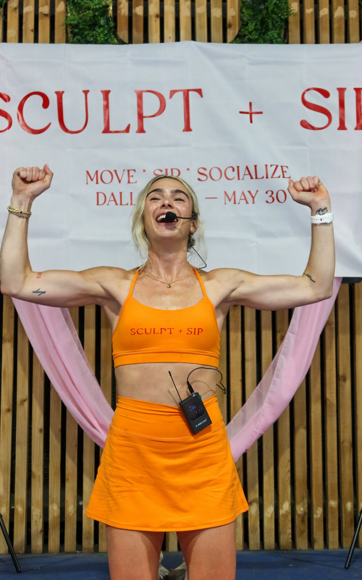 Woman in orange sportswear flexing arms, smiling, in front of a banner with text, outdoors with wooden background.