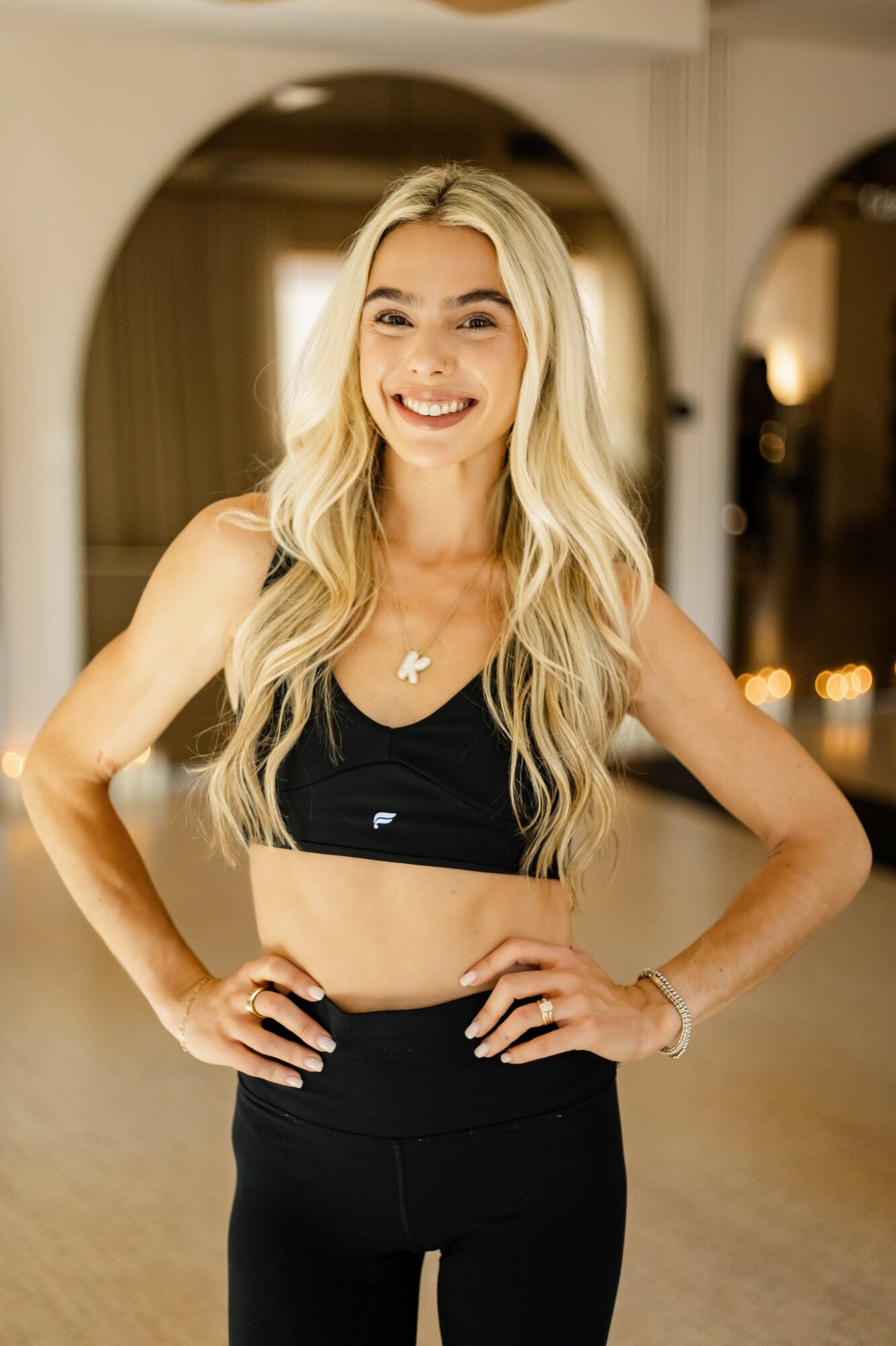 Smiling woman with long blonde hair in a black sports bra and leggings, hands on hips, indoors with arches and warm lighting