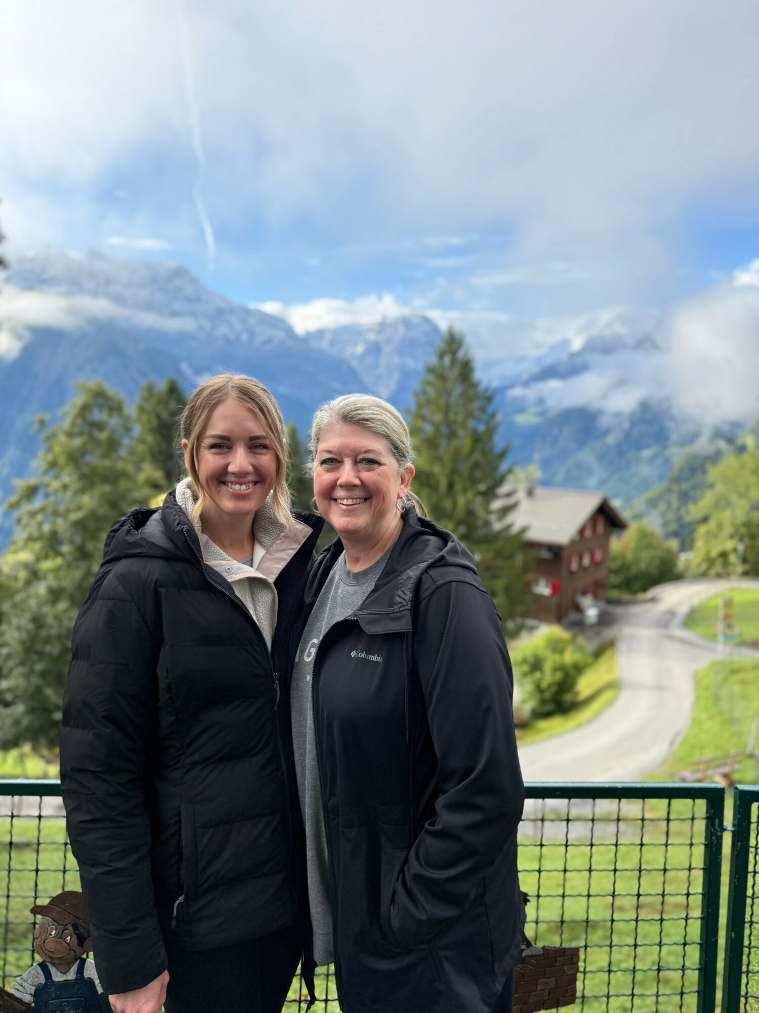 Two women stand outdoors with mountains and trees in the background, smiling and wearing black jackets.