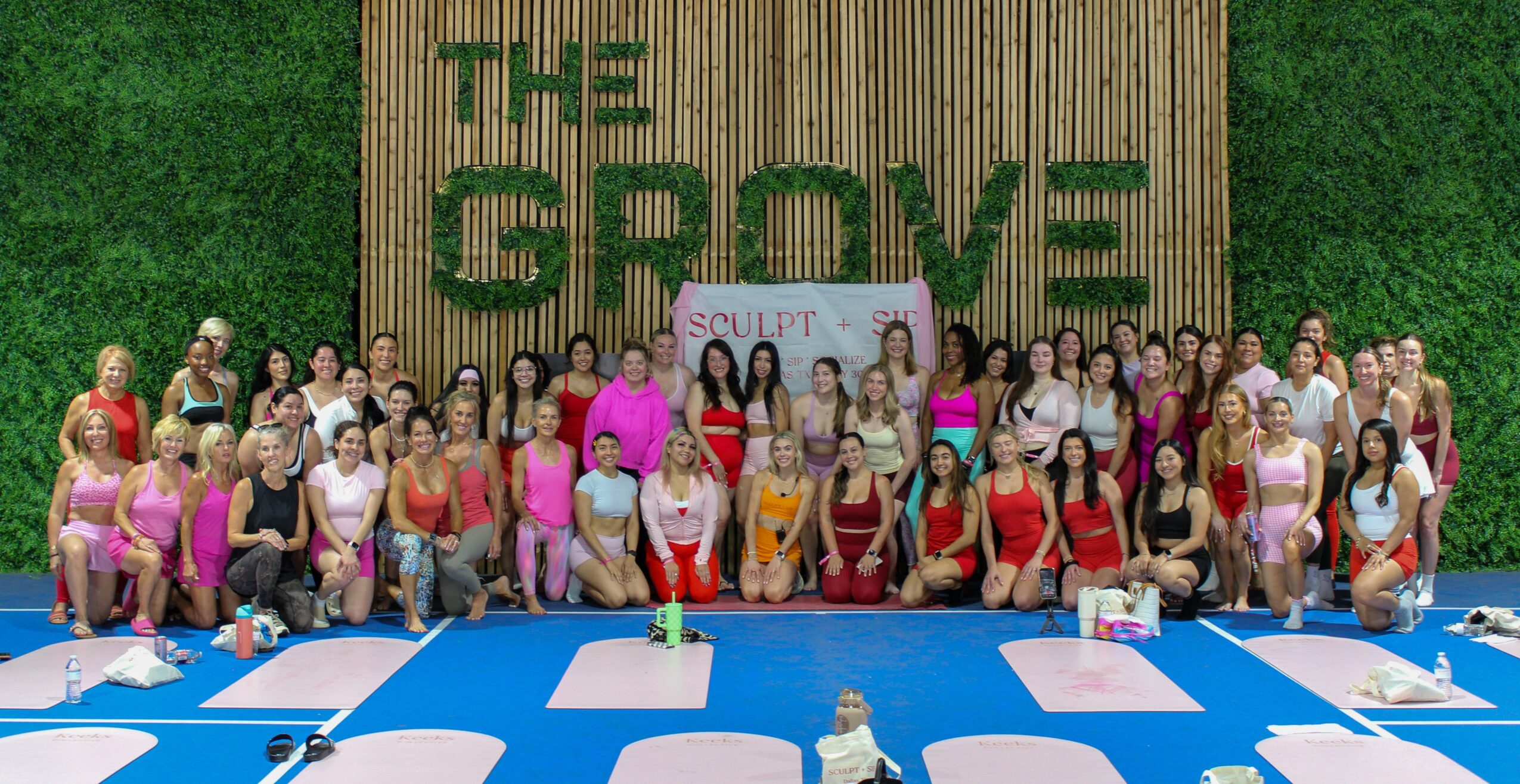 Group of people gathered outdoors in front of a large wooden sign reading 'THE GROVE' with green grass background.