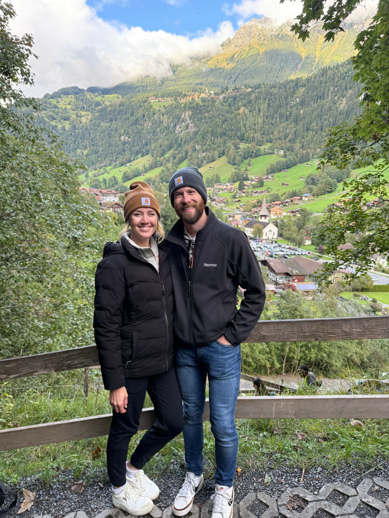 Two people standing outdoors on a wooden bench, surrounded by green trees and hills, with a village in the background.