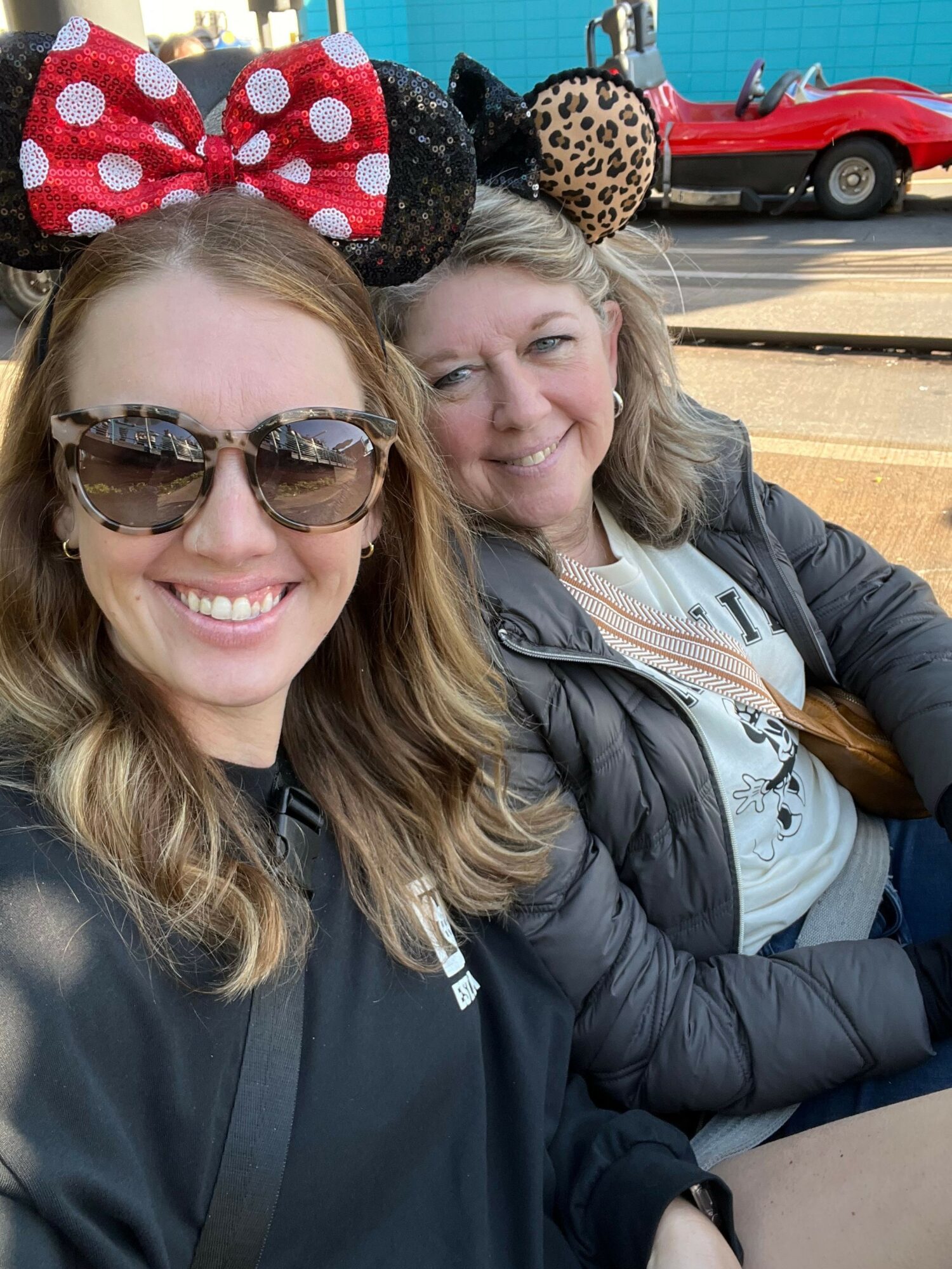 Two women smiling outdoors, one wearing sunglasses and a large polka-dot bow headband, the other with long hair and a jacket.