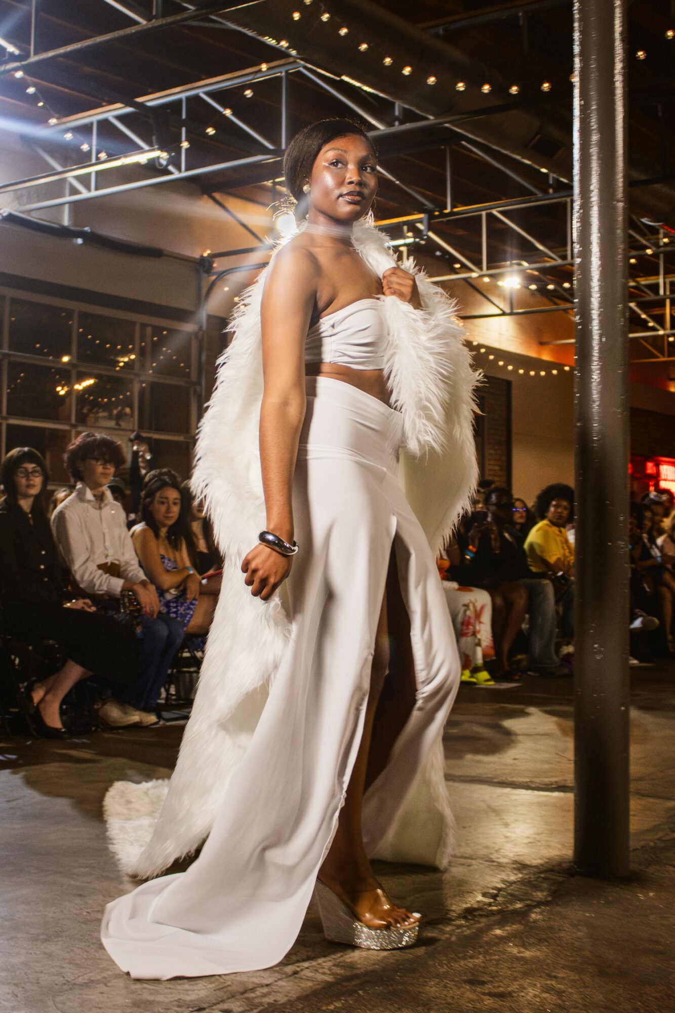 Woman in white strapless top and wide-leg pants with feathered accessory, standing indoors at night, audience in background.