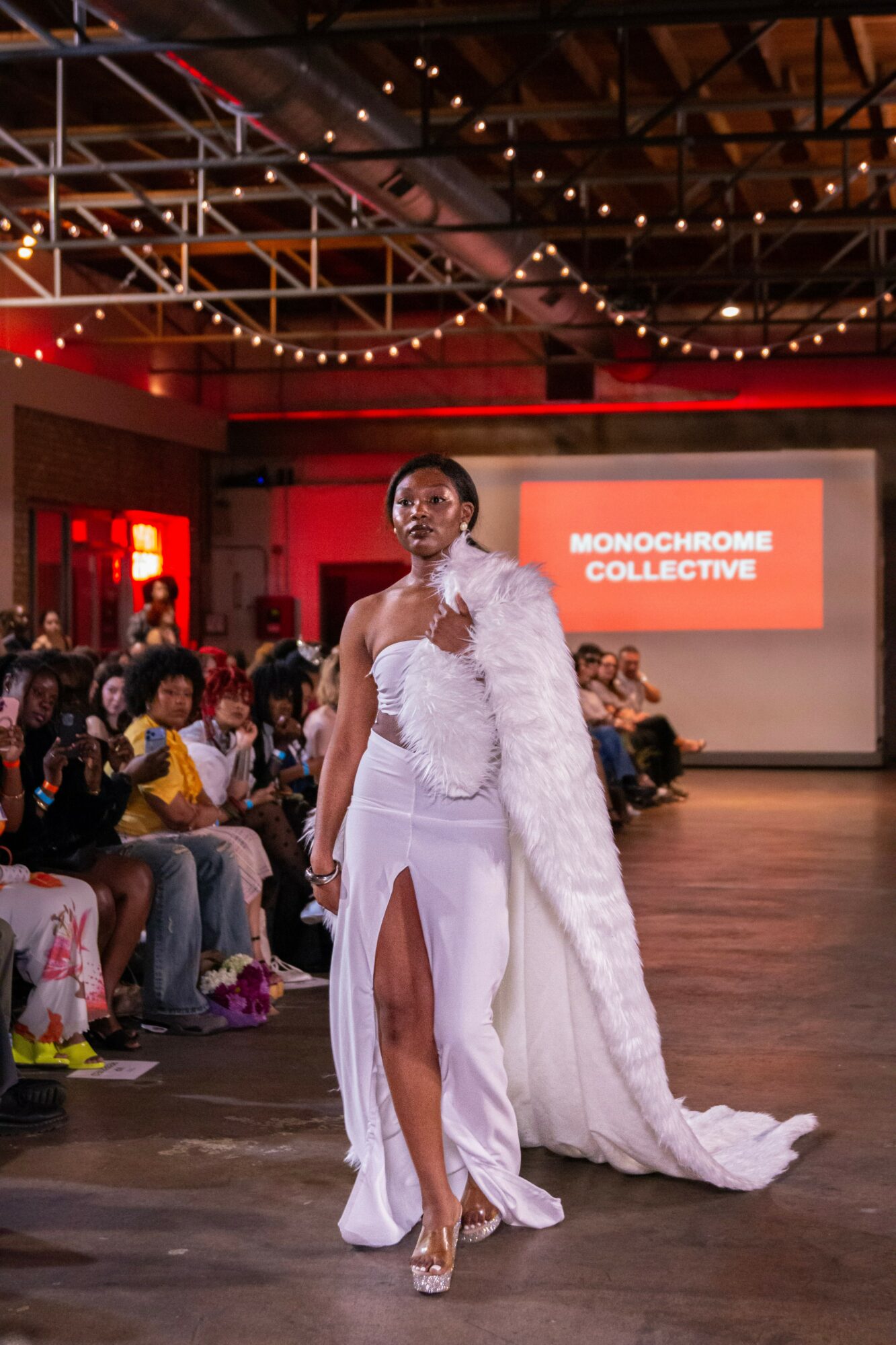 Model walking on runway in white dress with high slit, audience seated on sides, industrial ceiling with string lights, screen displaying 'Monochrome Collective'.