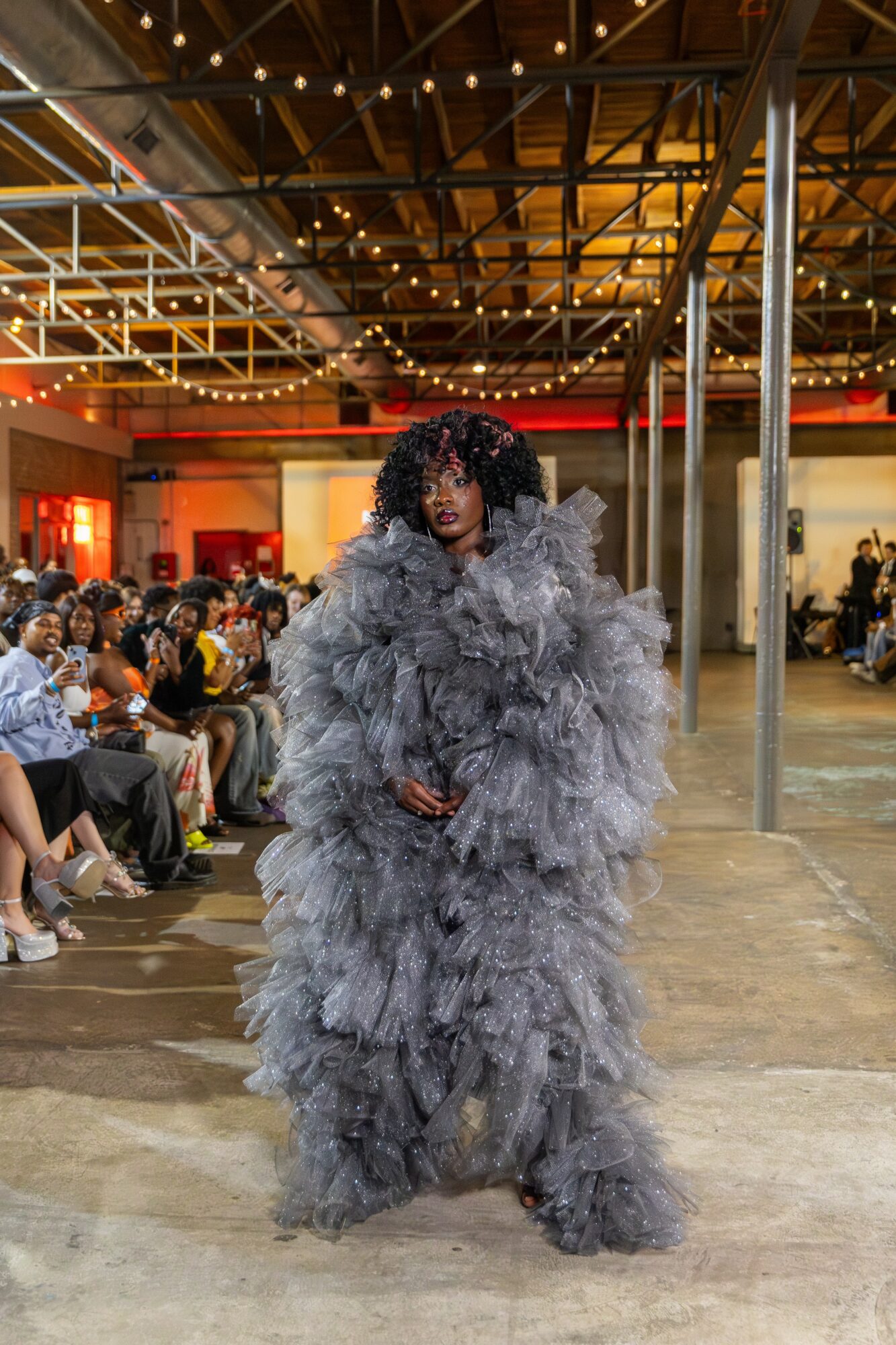 Person in a large, fluffy gray dress with curly hair walking in an indoor event space with seated audience.