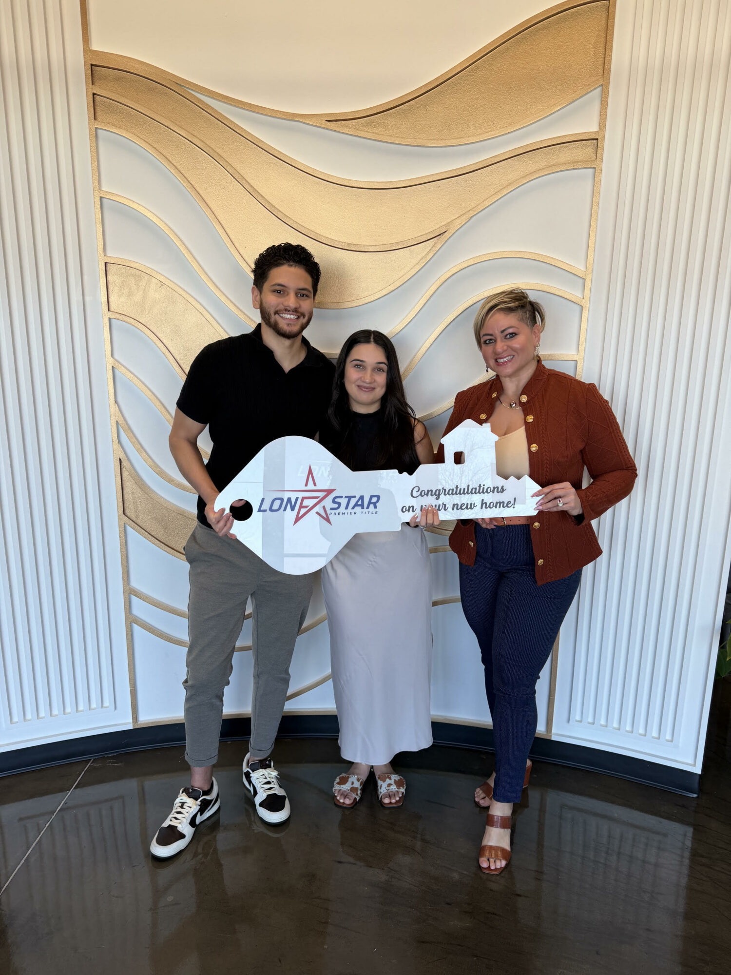 Three people standing together indoors, holding a large check and a certificate, smiling in front of a decorative wall.