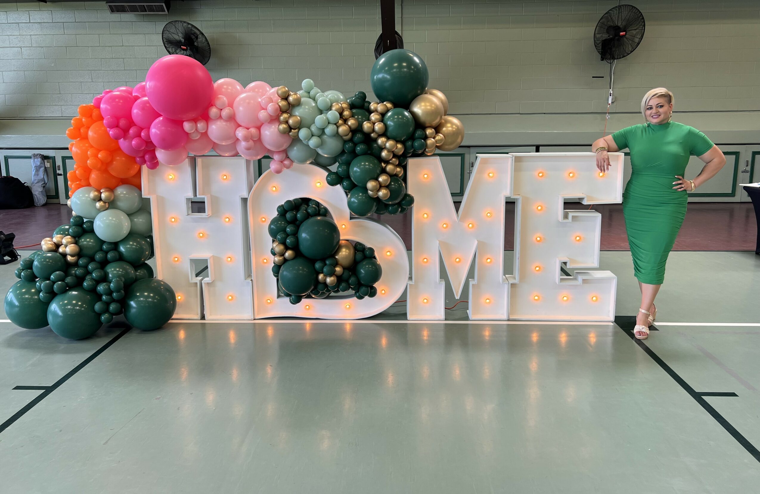 Large illuminated letters spell 'HOME' with colorful balloons arranged around them, and a woman in a green dress standing beside.