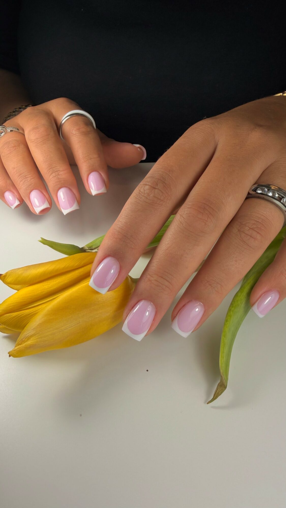 Two hands with rings resting on a white surface, near yellow flowers and green leaves, with pink nail polish.